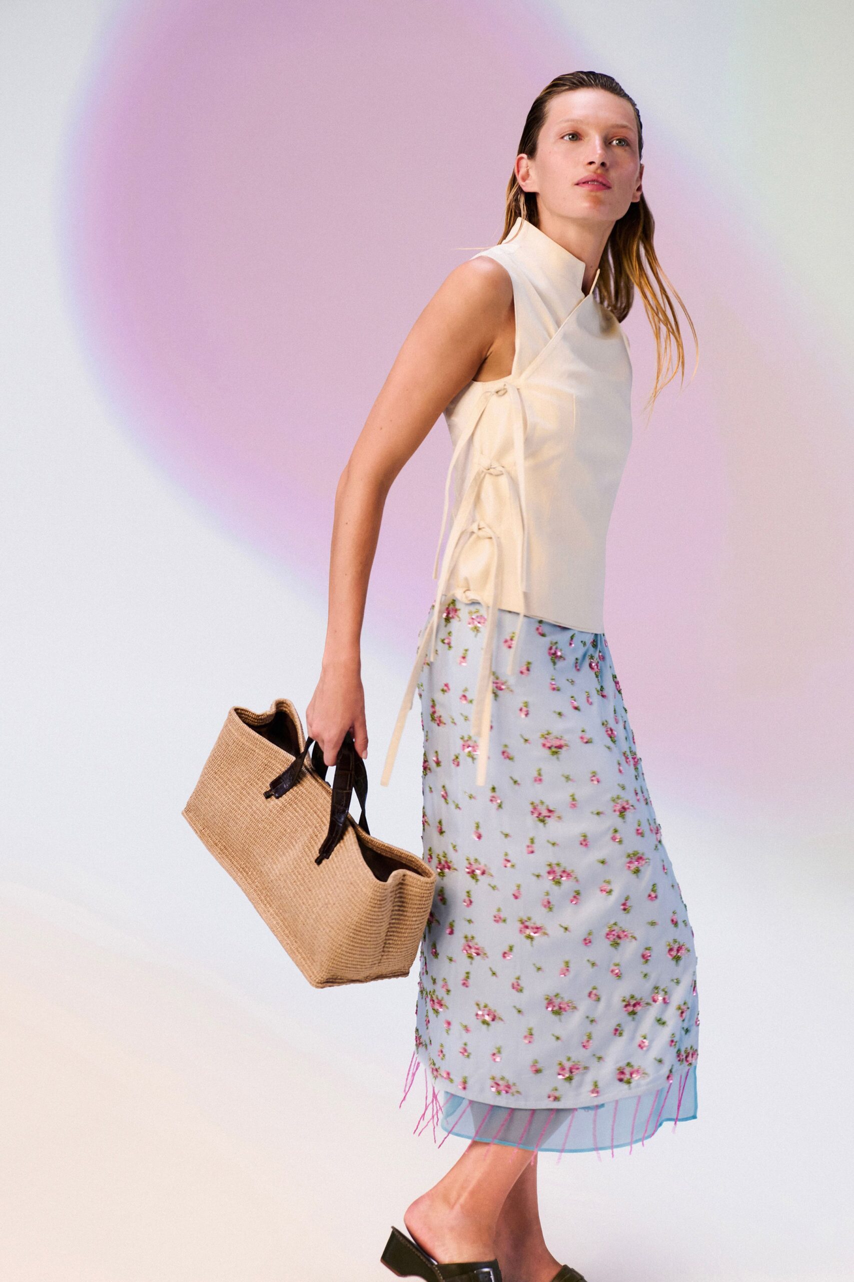 Cream sleeveless wrap top with frog closures climbing the side seam. Powder-blue floral slip skirt, pink thread fringe flicking below the hem. Straw tote with dark leather handles folding inward at the rim. Black square-toe mules with low block heels. Carrying peaches back from the farm stand before the heat breaks.