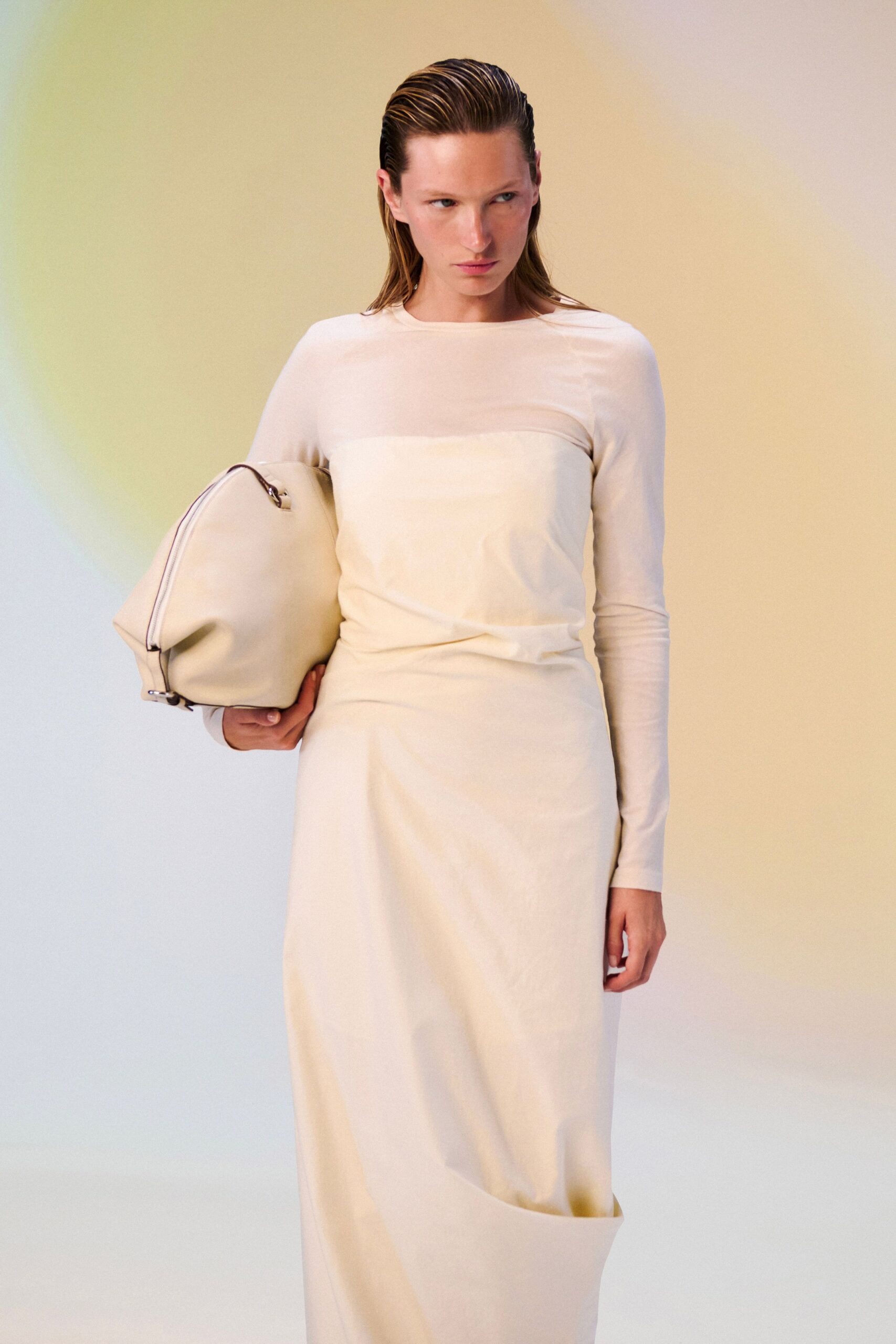Ivory column dress with a sheer yoke and long close sleeves. Oversized cream holdall in smooth leather, dark piping tracing the zip. Early hotel checkout, carrying more than fit back in the suitcase.