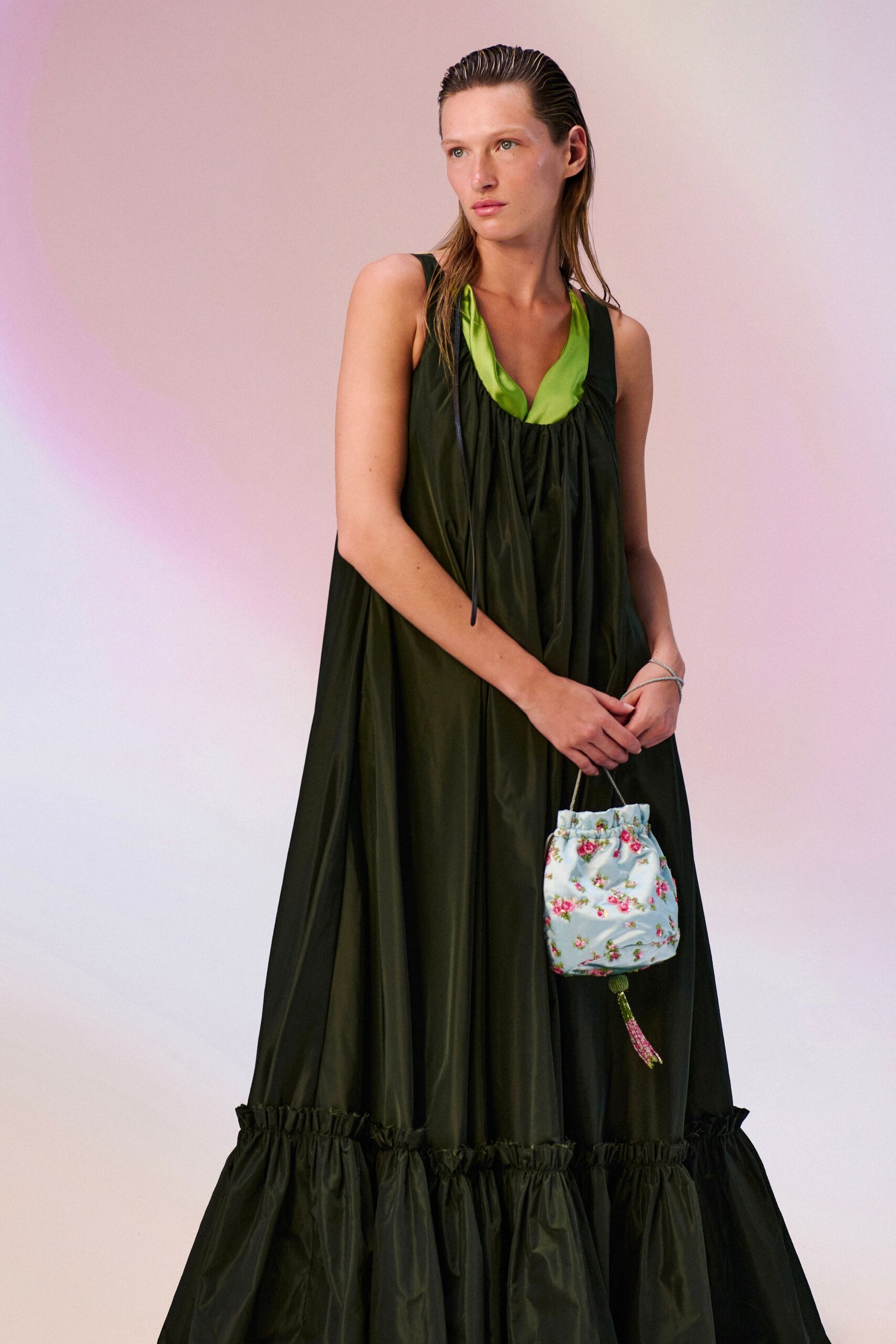 Black technical-satin tent dress, gathered scoop neck over a lime inset. Tiered ruffle hem rustling wide around the ankles. Thin silver cords looped twice at the wrist. Powder-blue floral pouch with a beaded tassel hanging low. Walking back from the hotel pool with dinner shoes still in mind.