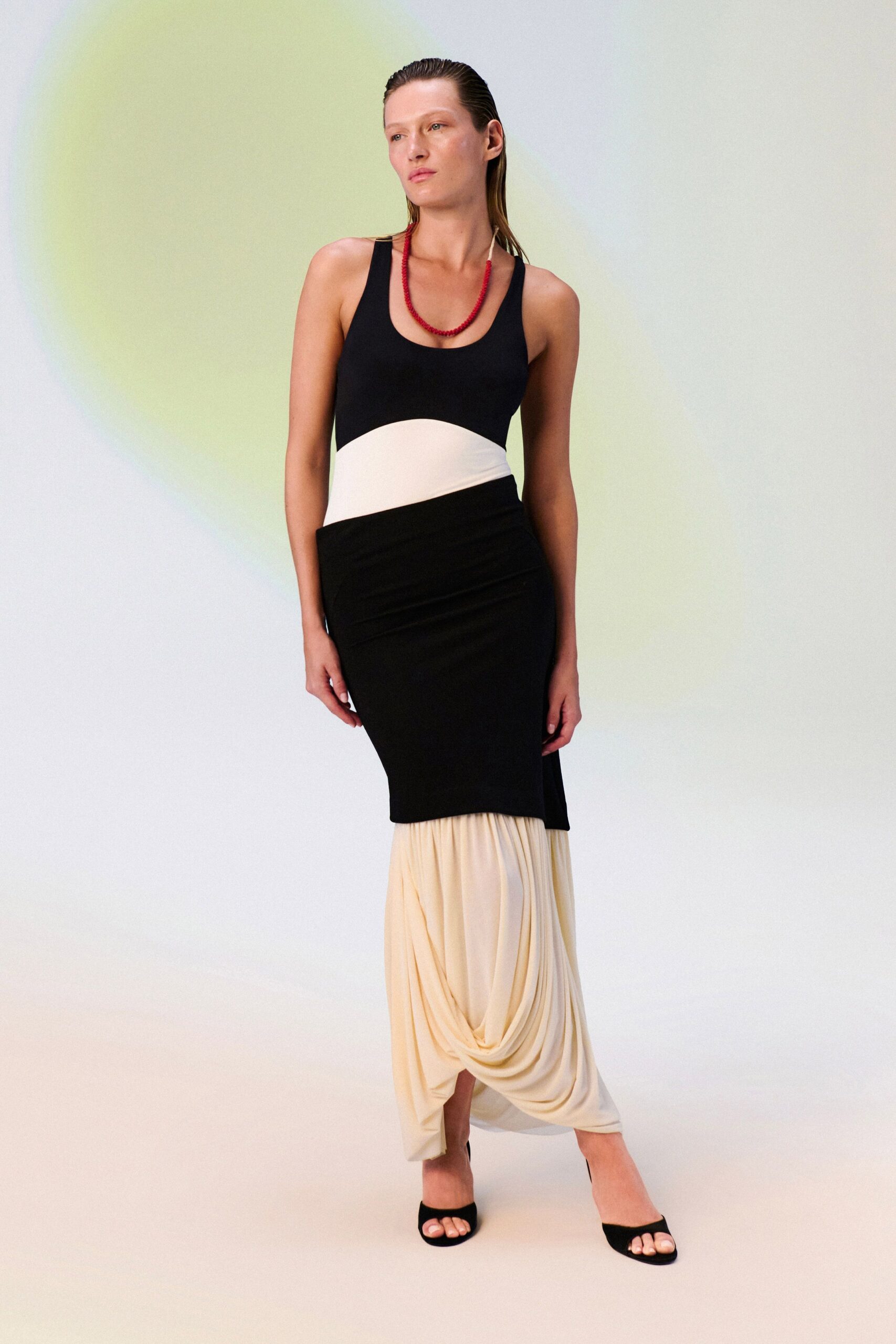Short strand of tiny red beads, slightly uneven at the clasp. Black jersey tank dress cut with a curved cream waist panel and pooled draped underlayer. Black suede slide mules with a squared open toe. For drinks on a hotel terrace while the umbrellas are still folded.
