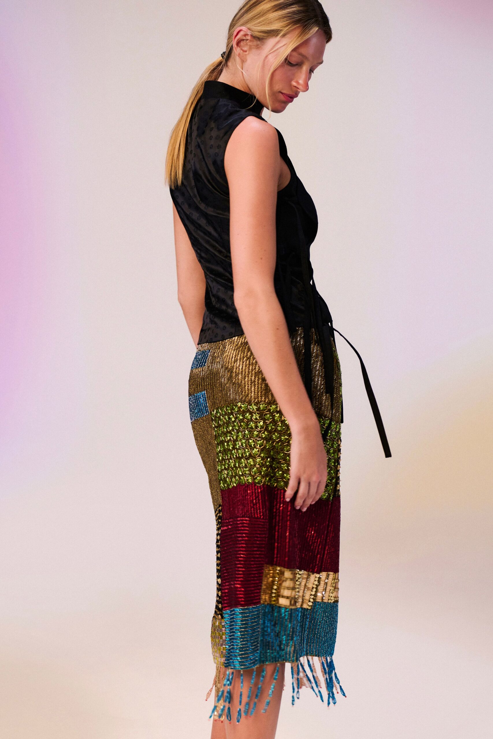 Black jacquard shell top, side lacing hanging loose in long cords. Patchwork beaded skirt in metallic blocks, blue fringe brushing the knee. Hotel bar carpet after midnight, still carrying the conference badge crease.