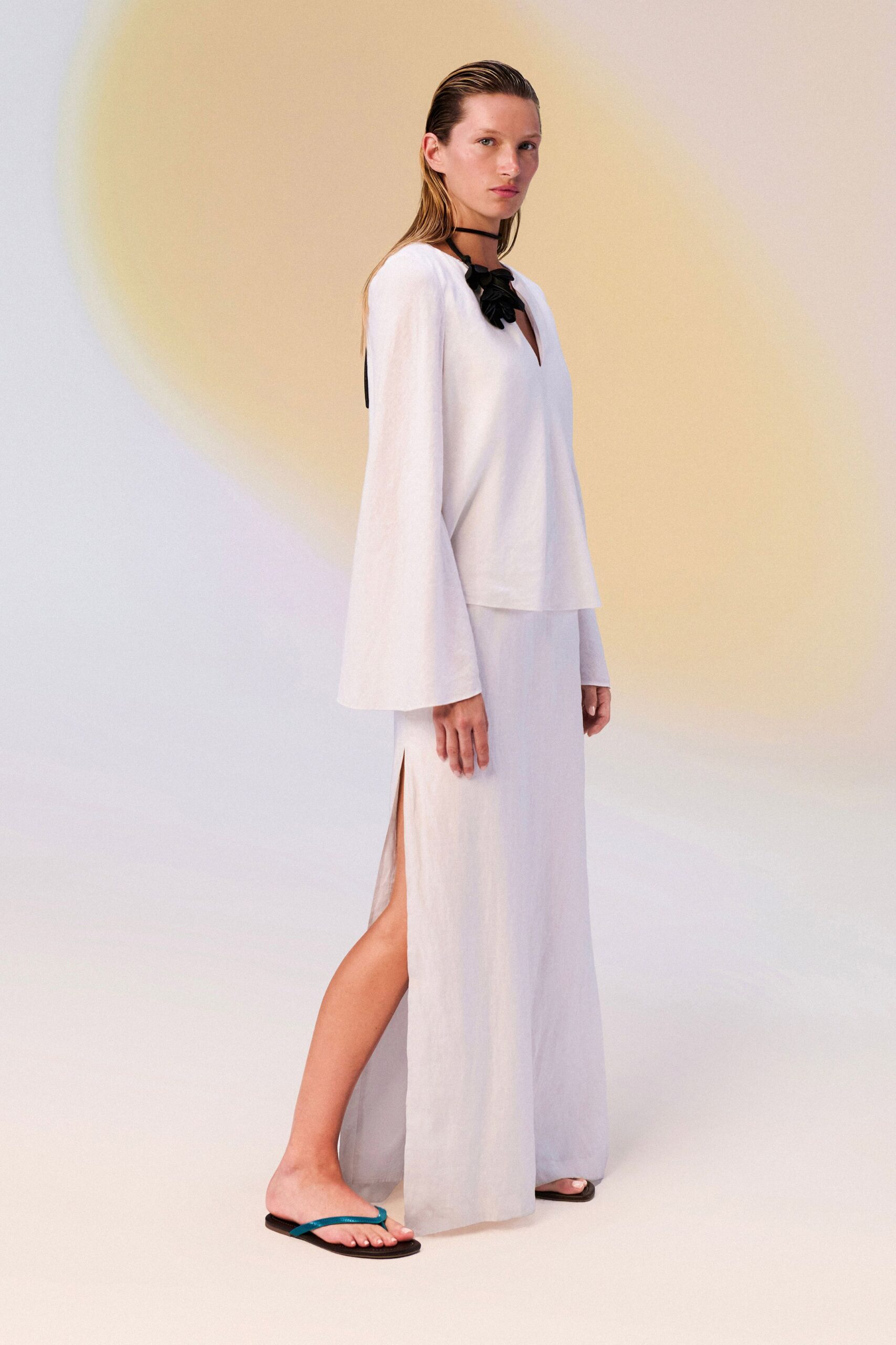 White linen tunic with a deep notch neck and bell sleeves. Black cord tie knotted with clustered petal-like tabs at the throat. Column skirt in the same slubbed weave, slit high at one side. Black flip-flops with bright turquoise thong straps. Walking barefoot to rinse salt off at the outdoor shower.