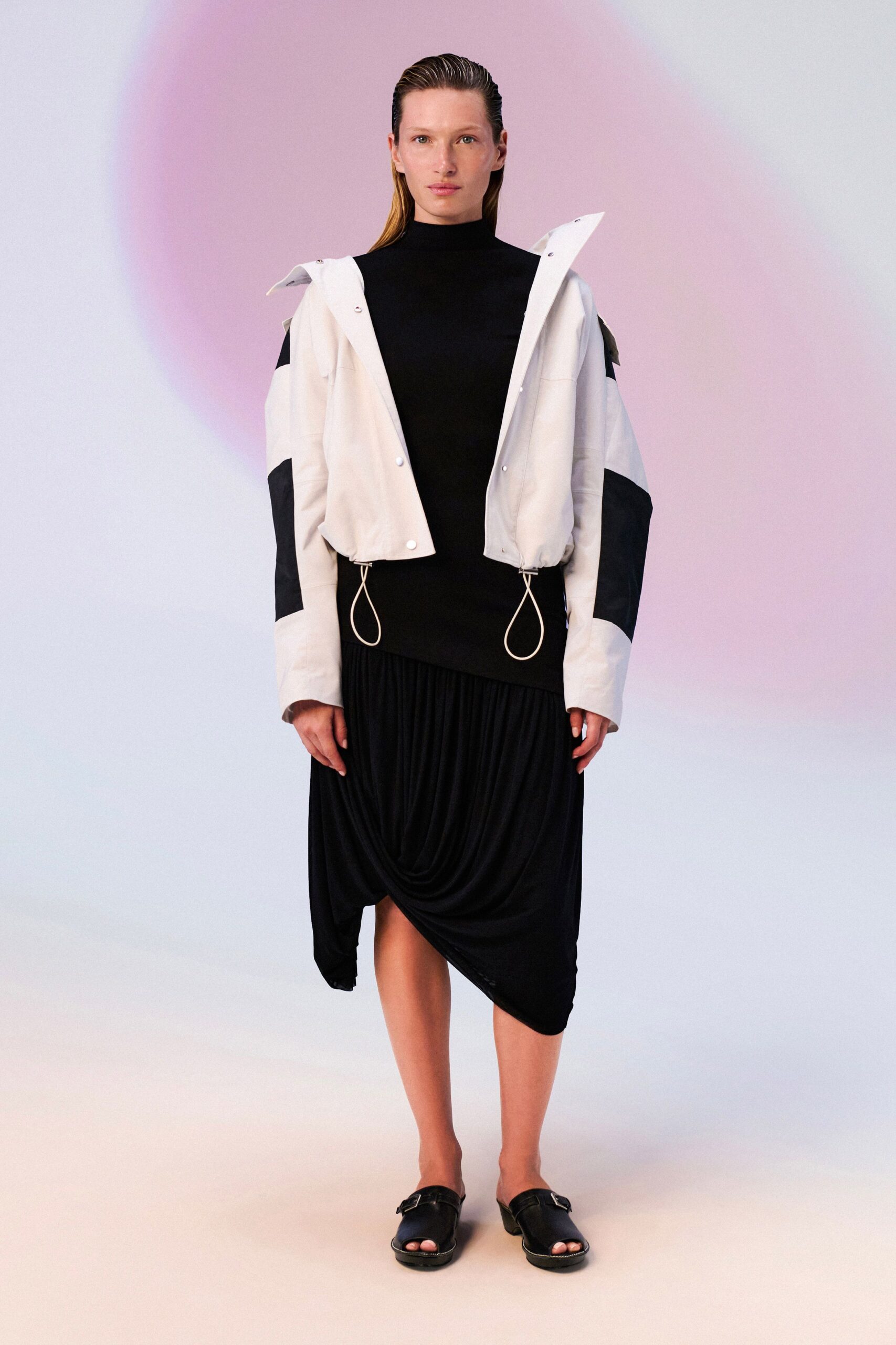 White snap-front jacket with black sleeve panels and dangling drawcord loops. Black mock-neck dress, jersey draped into a low cowl across the skirt. Black leather mule sandals with a square silver buckle. Walking from the ferry into a wind that turns warm by noon.