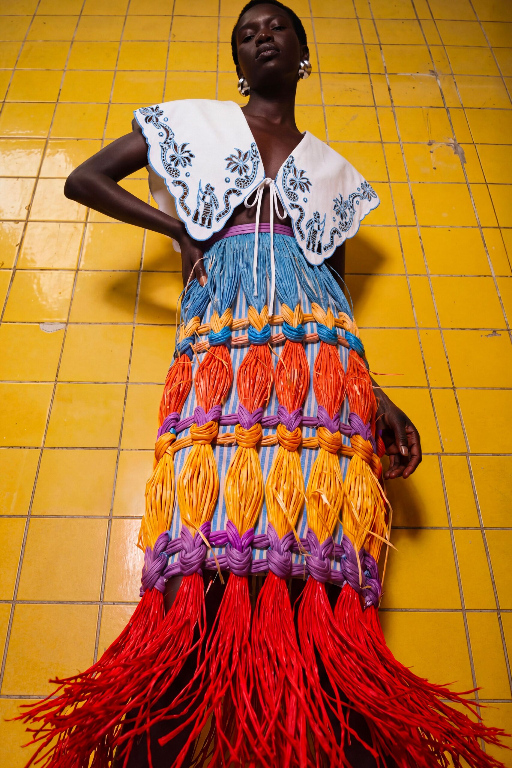 White capelet with scalloped cutwork and blue embroidery, tied open at the bust. Blue striped skirt caged in thick braided cords, ending in long red raffia fringe. Chunky silver hoops. Walking past shuttered fruit stalls after the pavement has finally cooled.