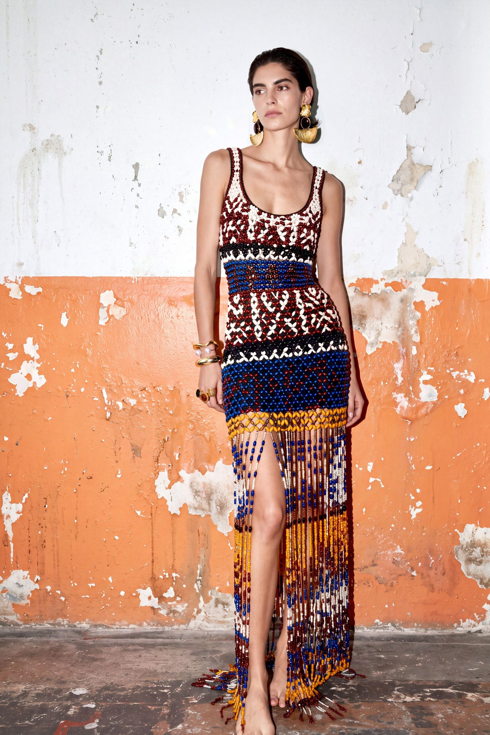 Hammered gold drop earrings with dark oval links. Hand-beaded net dress in cream, oxblood, cobalt and marigold, fringe brushing the floor. Stacked gold cuffs with a single pearl caught between. Walking barefoot through a crumbling seaside villa after the power cuts out.