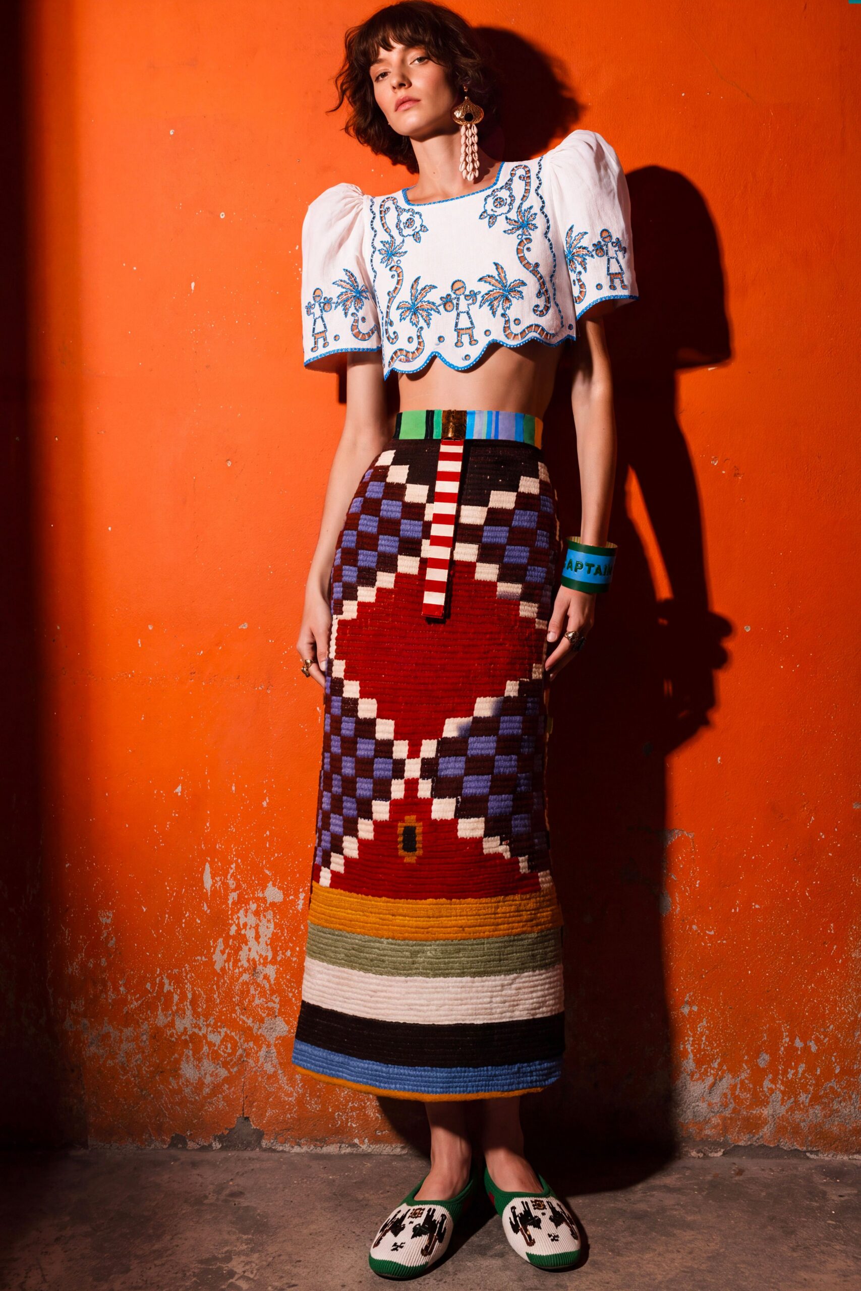 White cotton crop top with puff sleeves and blue folk embroidery. Tapestry-knit column skirt, checkerboard and heart motifs, striped hem weighty. Wide green cuff printed with block letters. Flat knit slippers with green trim and black figures. Afternoon in a tiled courtyard, wall heat still rising after lunch.