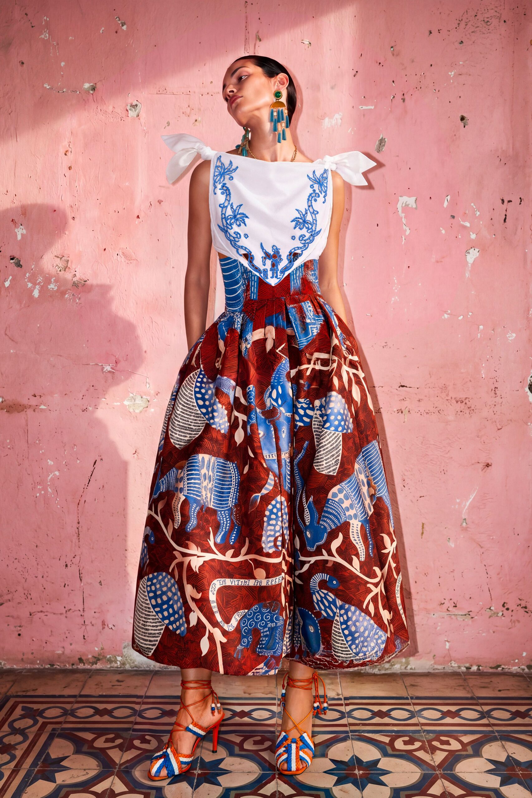 White cotton bib top with cobalt embroidery and tied shoulder bows. Voluminous red cotton culottes printed with blue armadillos and curling vines. Orange lace-up heels with braided blue-and-white bands across the toes. Long tassel drops in turquoise and amber. Courtyard wedding at noon, plaster walls throwing back the heat.