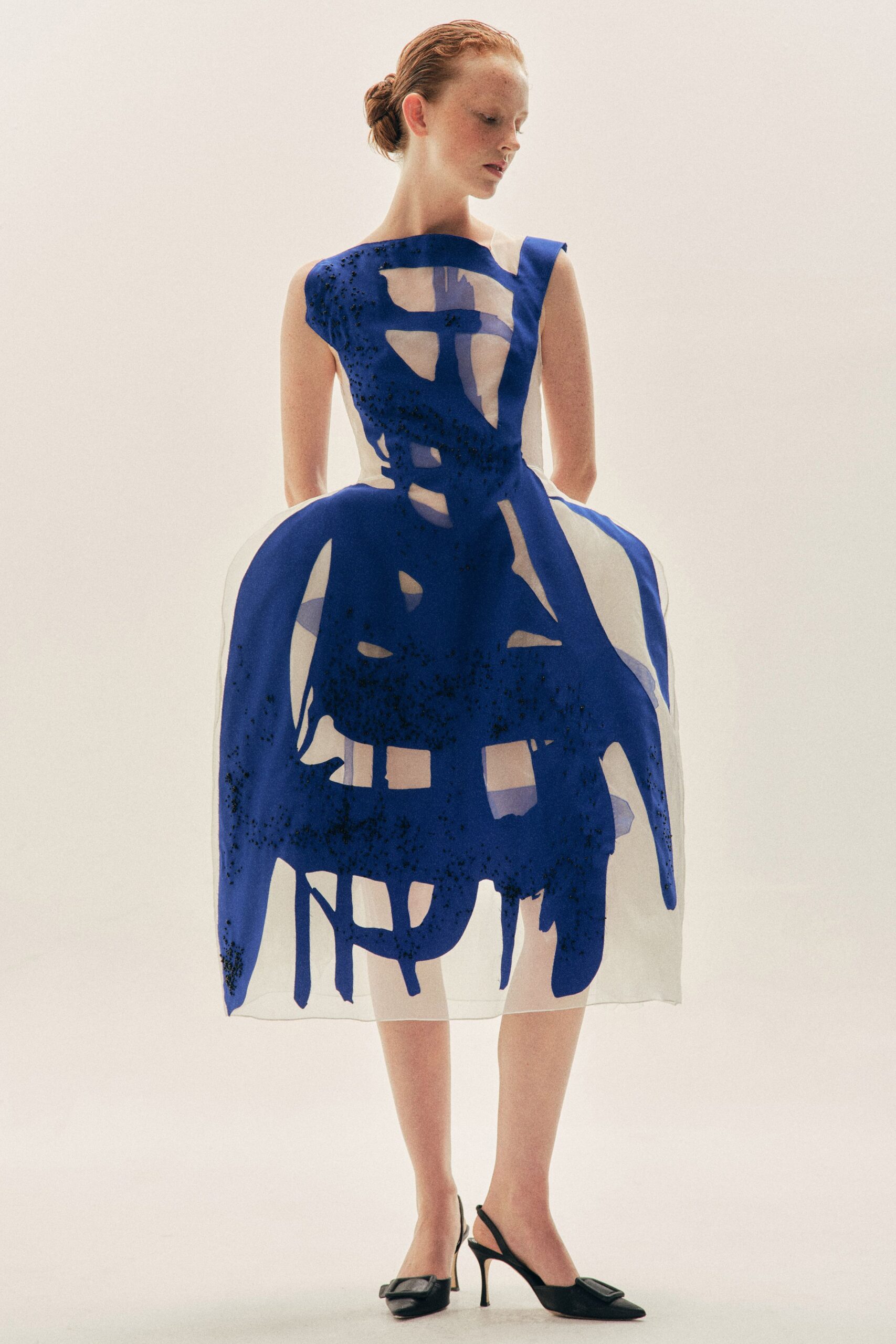Sheer organza dress with a cobalt painted overlay and scattered black beading. Black slingback pumps with oversized square buckles at the pointed toe. Museum atrium at noon, the floor still smelling faintly of mop water.
