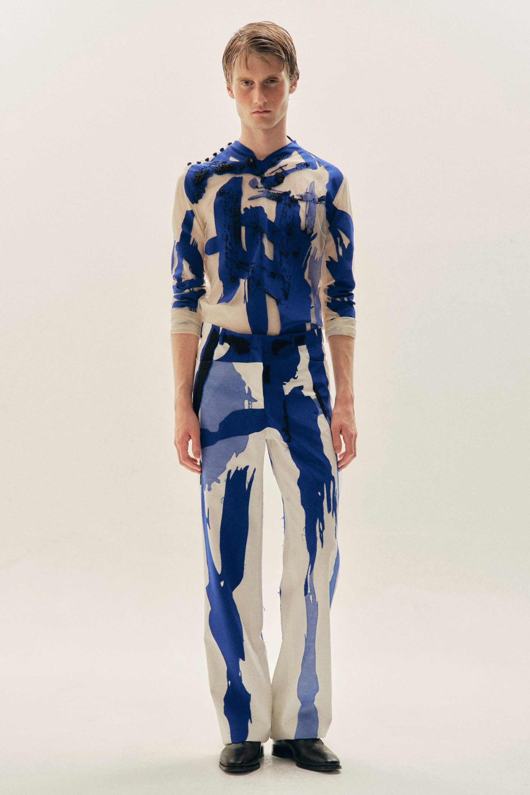 Cream knit top streaked with cobalt brushstrokes and knotted beaded trim. Matching straight-leg trousers, print dropping in long drips past the knee. Black square-toe boots. Museum loading dock just after the paint crew has gone home.