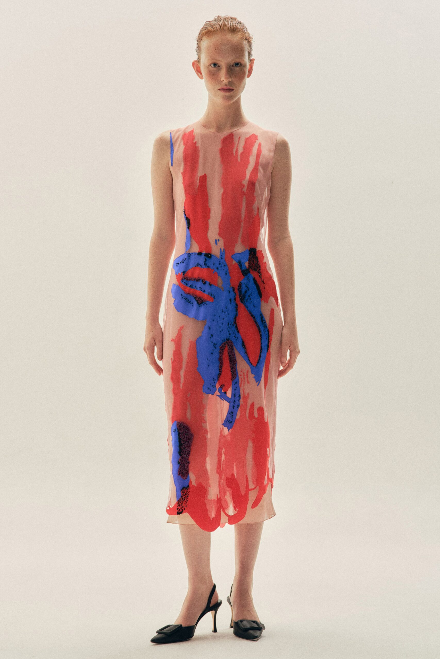 Sheer shell-pink sleeveless column, overprinted with scarlet strokes and cobalt bloom shapes. Black slingback pumps with square buckles and pin-thin heels. Museum atrium at opening hour, flowers arriving before the guests.