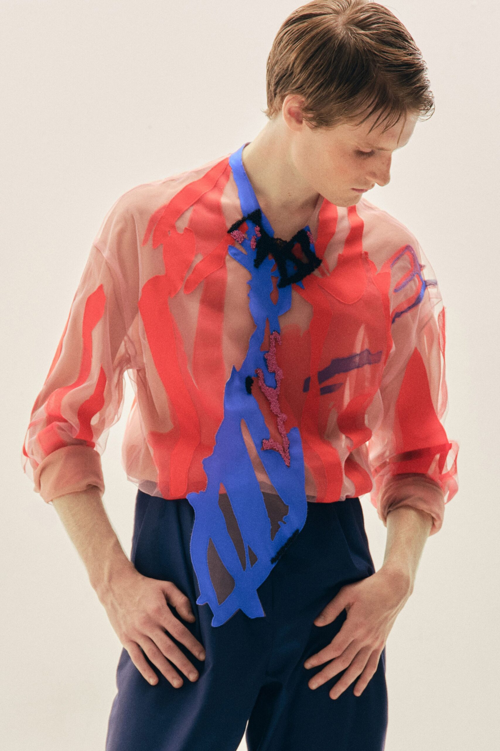 Sheer blush organza blouse, washed with coral and cobalt cutout appliques. Deep blue trousers falling plain beneath the crowded translucent top. Afternoon light in an art school hallway, poster paint still tacky.