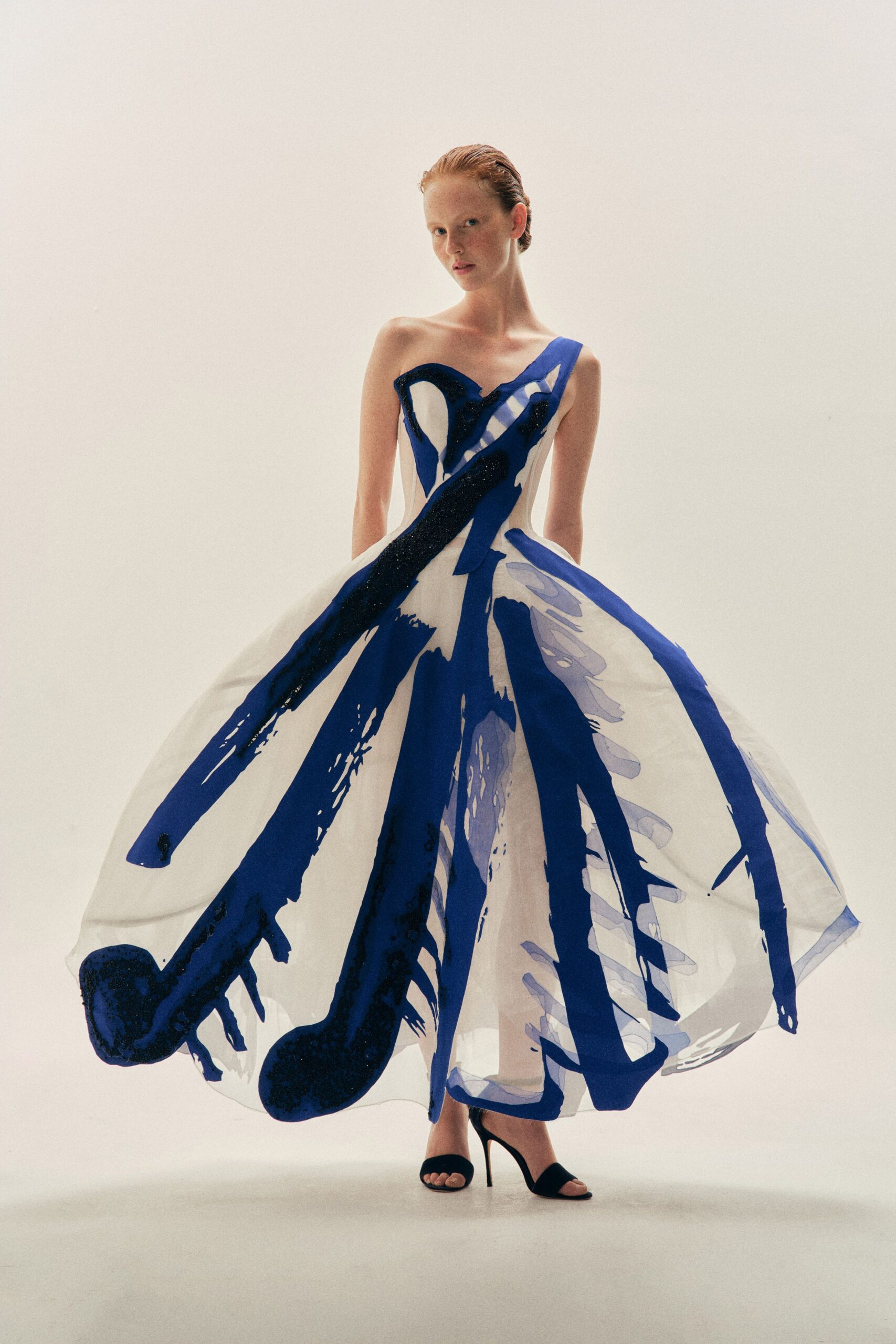 One-shouldered white ball gown with cobalt brushstroke panels over a sheer underlayer. Black suede mule sandals with a slim stiletto heel. Gallery atrium at closing, still carrying the scale of the paintings.
