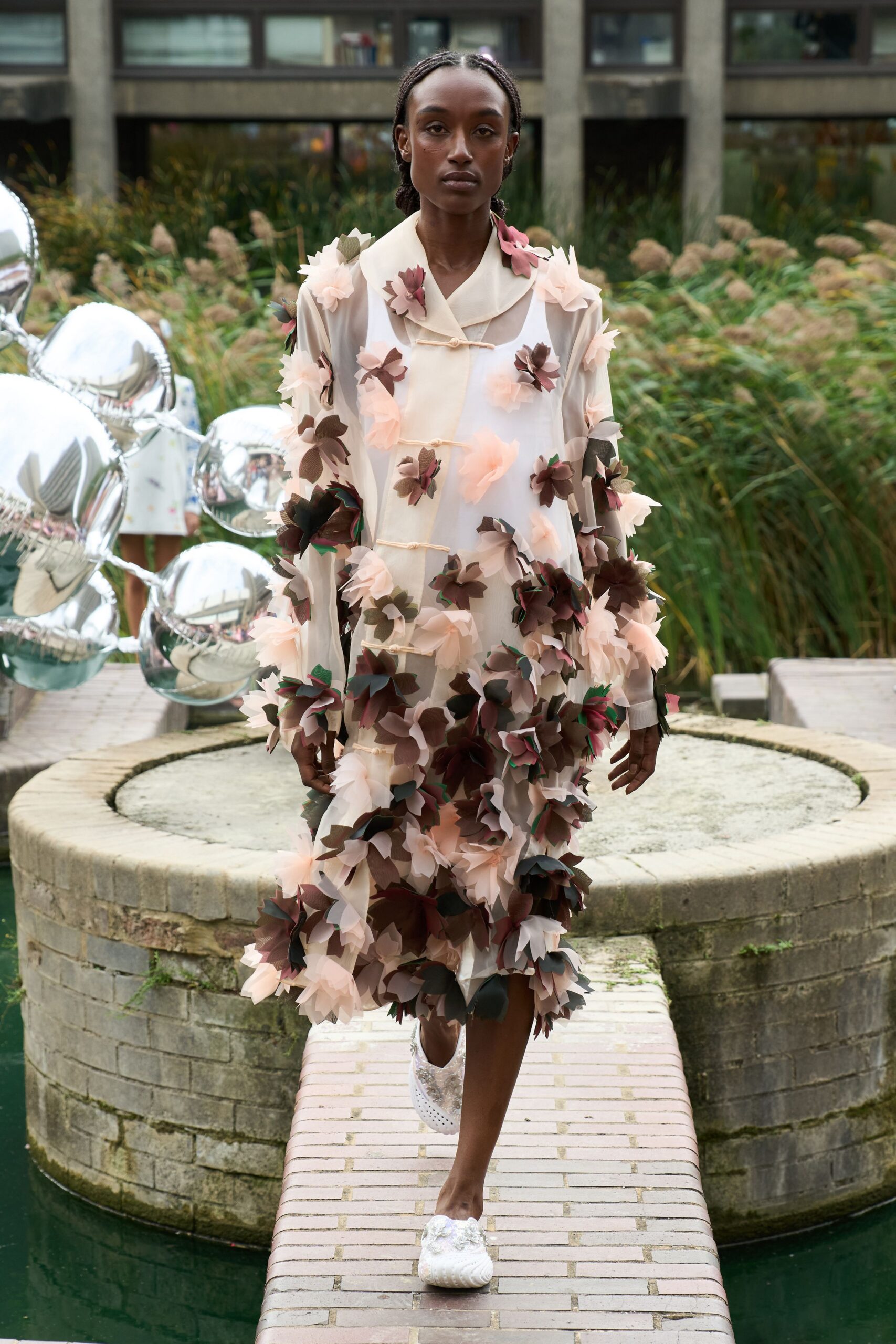 Sheer ivory organza coat dress with frog closures and appliqued chiffon flowers thickening at the hem. Opaque white slip glowing beneath the translucent layers. White brocade flats with petaled trim at the vamp. Crossing the garden bridge while the fountain's been switched off for winter.