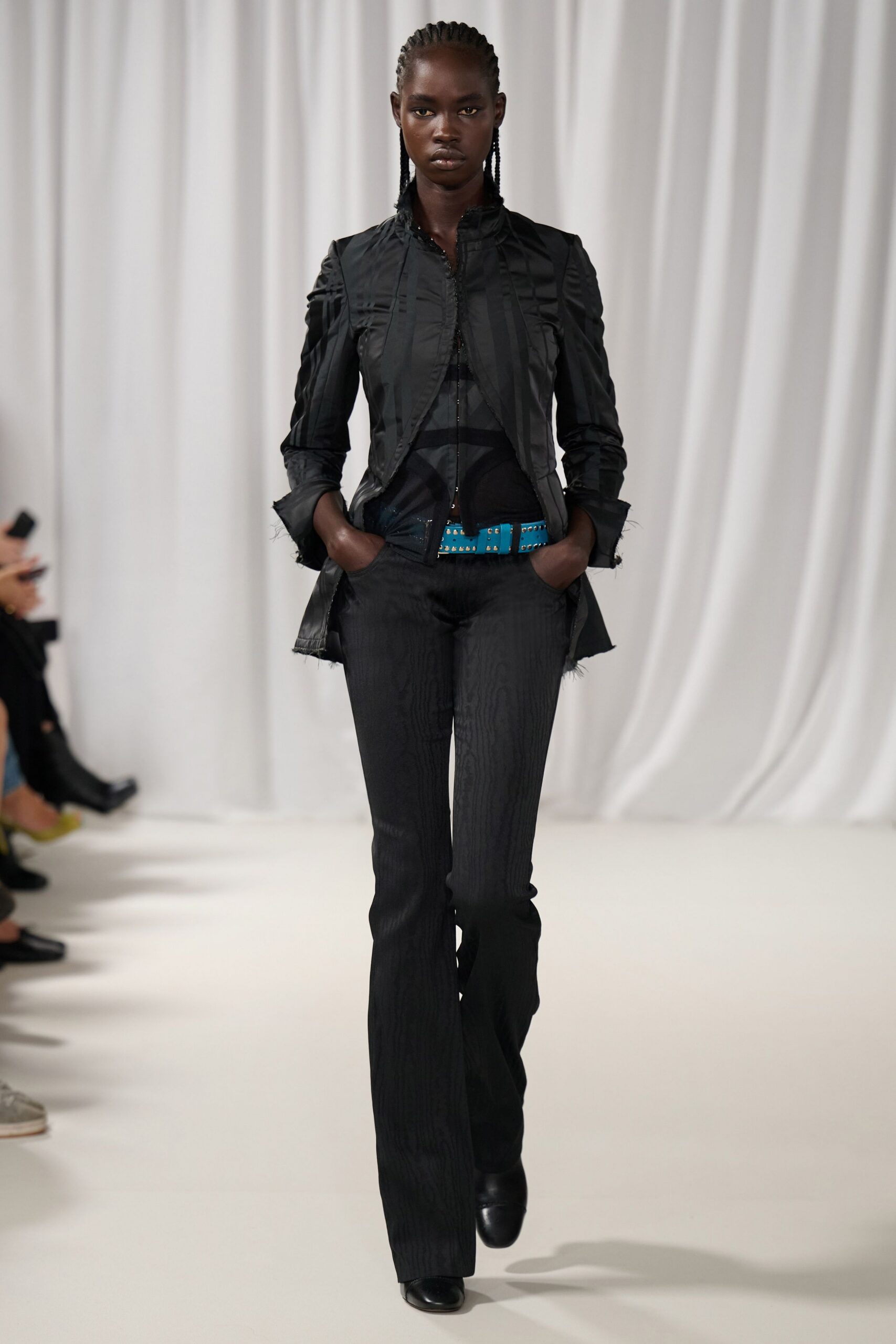 Black fitted jacket with puckered seams and frayed cuffs. Sheer black mesh top, seamed into curved opaque panels. Turquoise studded belt cutting bright across the waist. Black trousers with a faint moire stripe and slight flare. Square-toe black leather boots. Walking out of a matinee into cold sun and traffic spray.