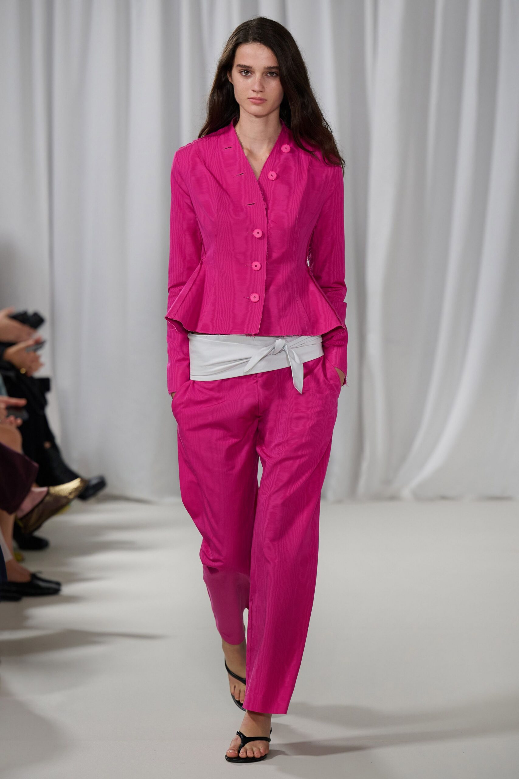 Hot-pink moire jacket, curved peplum kicked out at the hips. White wrap sash tied low, one end jutting sideways. Matching moire trousers cut straight and easy through the leg. Black thong sandals. For crossing a hotel lobby while the florist is still setting buckets out.