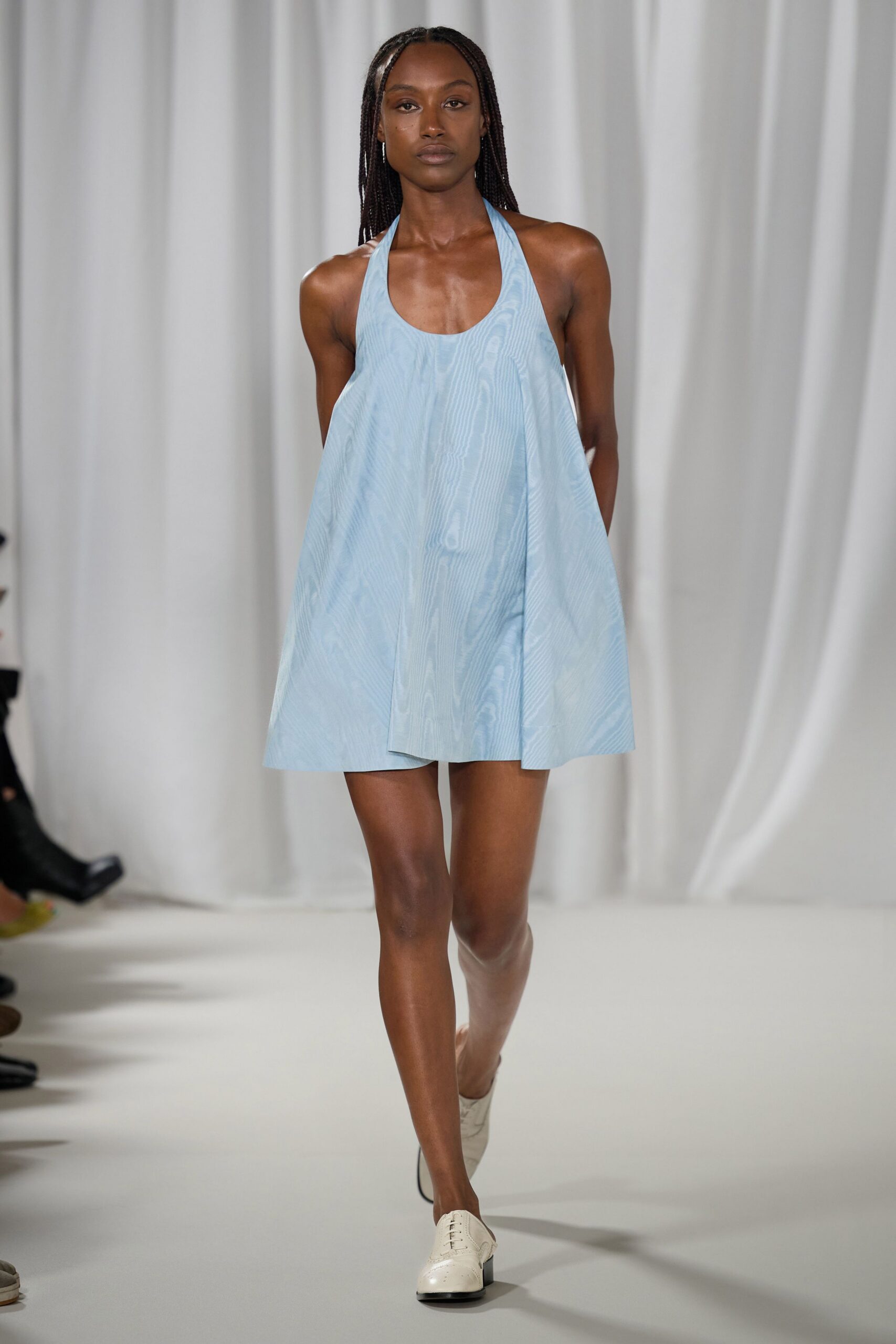 Powder-blue moire halter mini, cut in a stiff swinging trapeze. Cream leather oxfords with a dark stacked heel. Walking across a hotel ballroom before the chairs are set out.