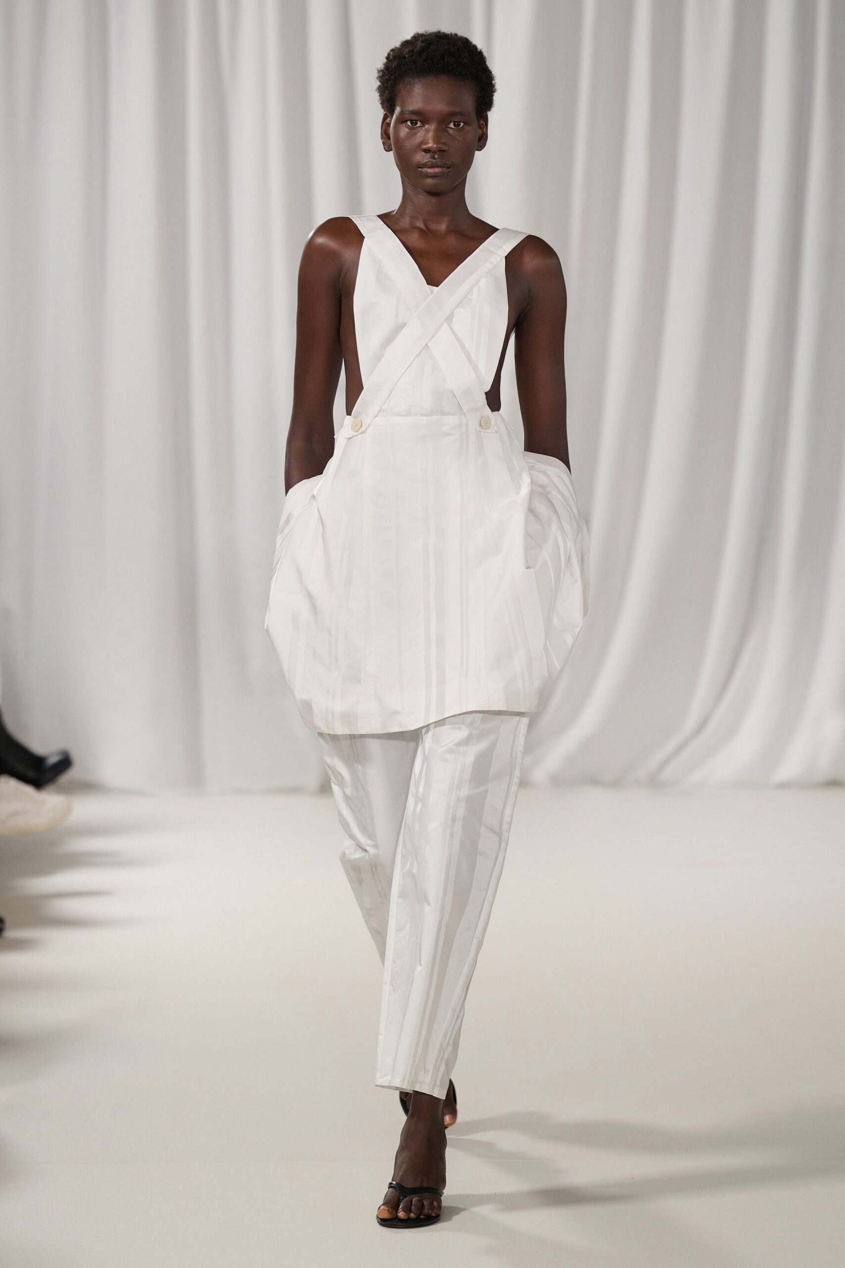 White cotton apron top with wide crossed straps and bare sides. Crisp white sleeves bunched loose at the forearms. White straight trousers with a pressed center crease and slight sheen. Black thong sandals. Walking across a sunstruck courtyard after changing out of kitchen whites.