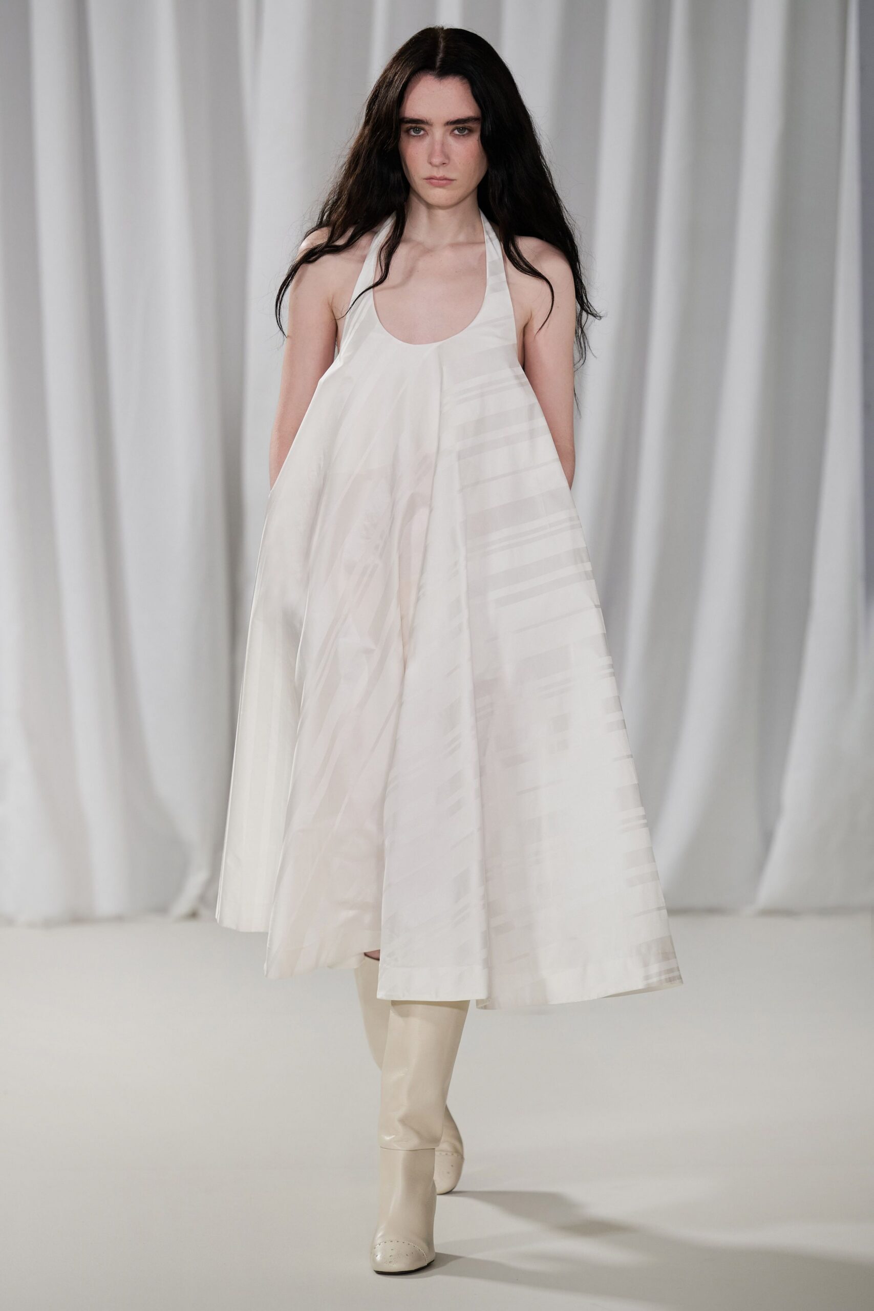 Milk-white halter dress in crisp woven cloth, flaring from a centered pleat. Cream knee boots with punched toe medallions and a blunt square front. Crossing a silent chapel hall after the flowers have been cleared.
