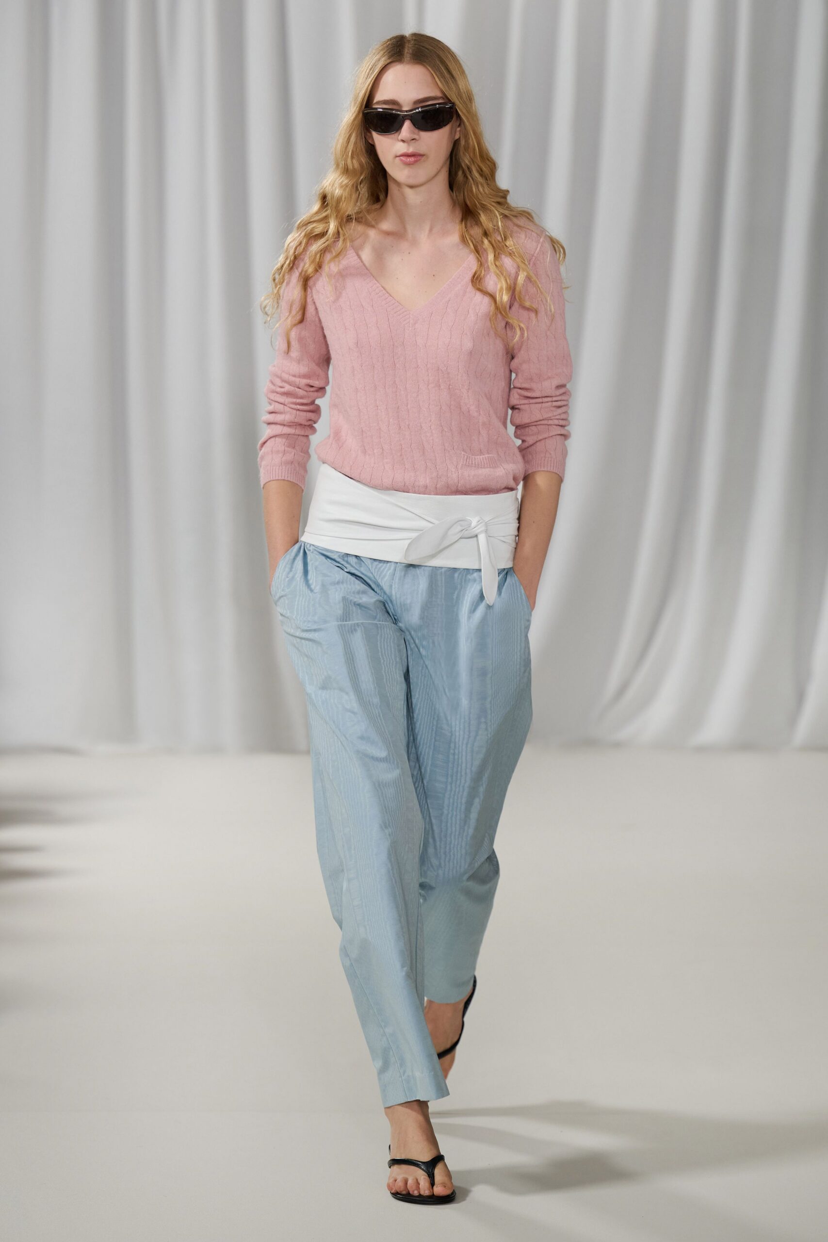 Narrow black wraparound sunglasses. Blush cable-knit V-neck sweater with a small patch pocket. Wide white sash tied off-center at the waist. Pale blue trousers in watery striped jacquard, cropped above the ankle. Black thong sandals. Walking the hotel terrace before breakfast, sea air still cool on your skin.