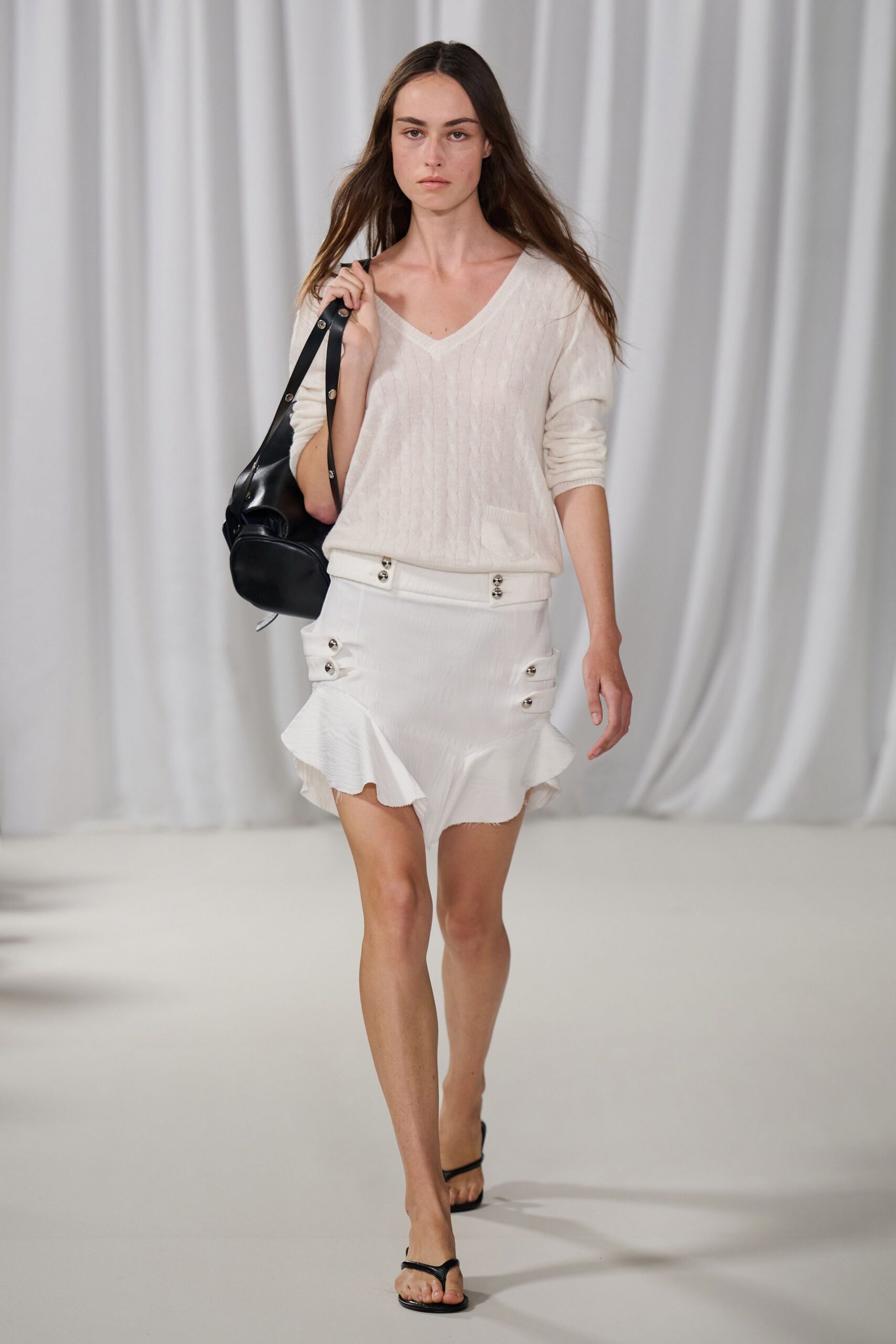 Ivory cable-knit V-neck sweater, slouched soft at the waistband. Black leather shoulder bag with silver rivets along the strap. Ivory ribbed mini skirt, flounced hem and side tabs fastened with twin studs. Black thong sandals. Walking from a beach hotel lobby before the towels have dried.
