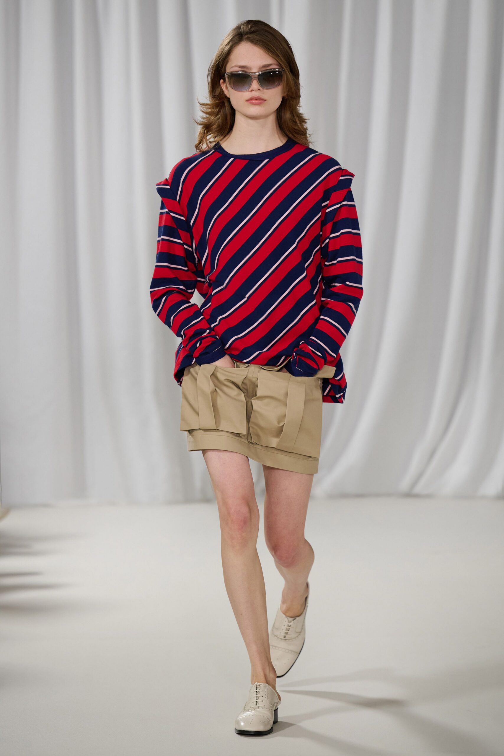 Square gradient sunglasses with a smoky translucent frame. Red and navy striped jersey top, shoulder tabs jutting out. Khaki cotton shorts with folded waistband panels hanging unevenly. Cream leather oxfords with punched brogueing at the toe. Walking to a ferry terminal with yesterday's shirt and a map folded in half.