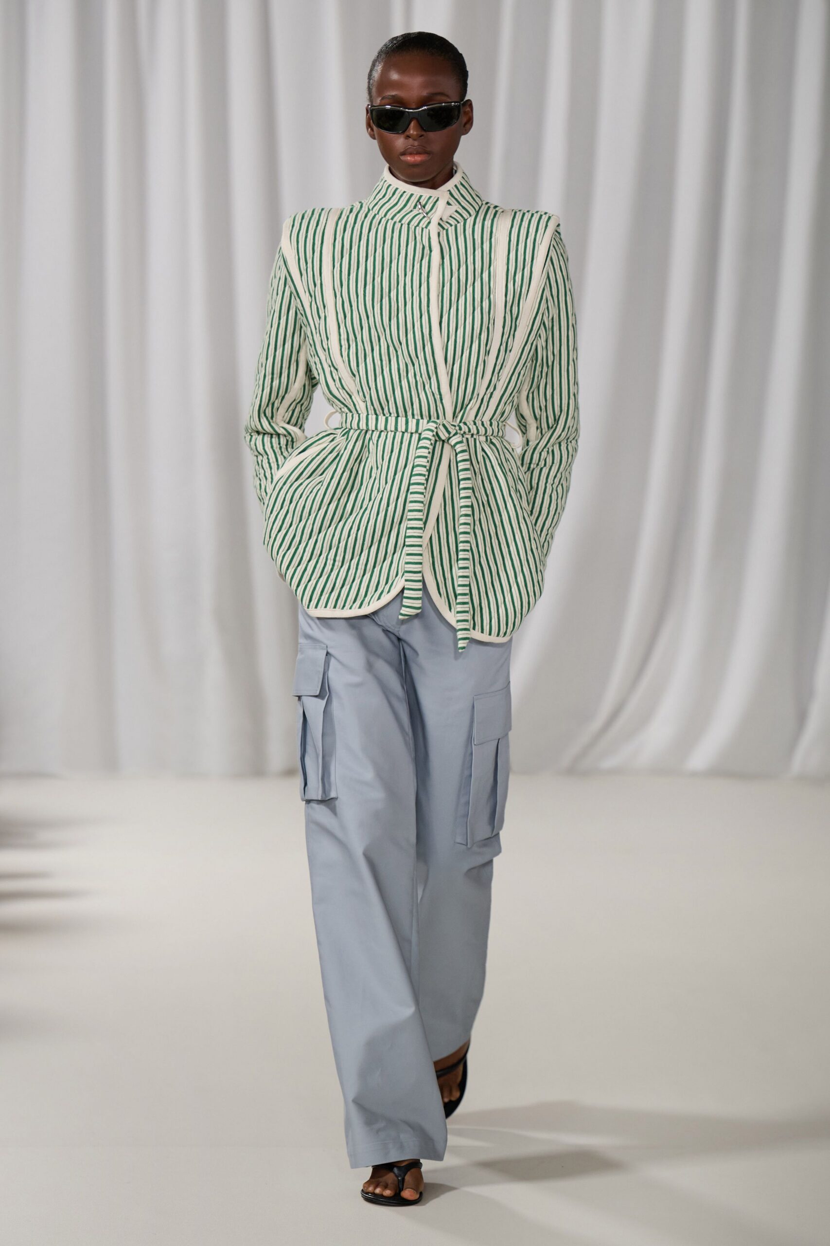 Narrow black wraparound sunglasses with a thin silver brow bar. Quilted ivory jacket traced with bottle-green stripes, tied askew at the waist. Pale blue cargo trousers with flat bellows pockets at each thigh. Black thong sandals, barely there. Walking the marina before noon, salt already drying on your arms.