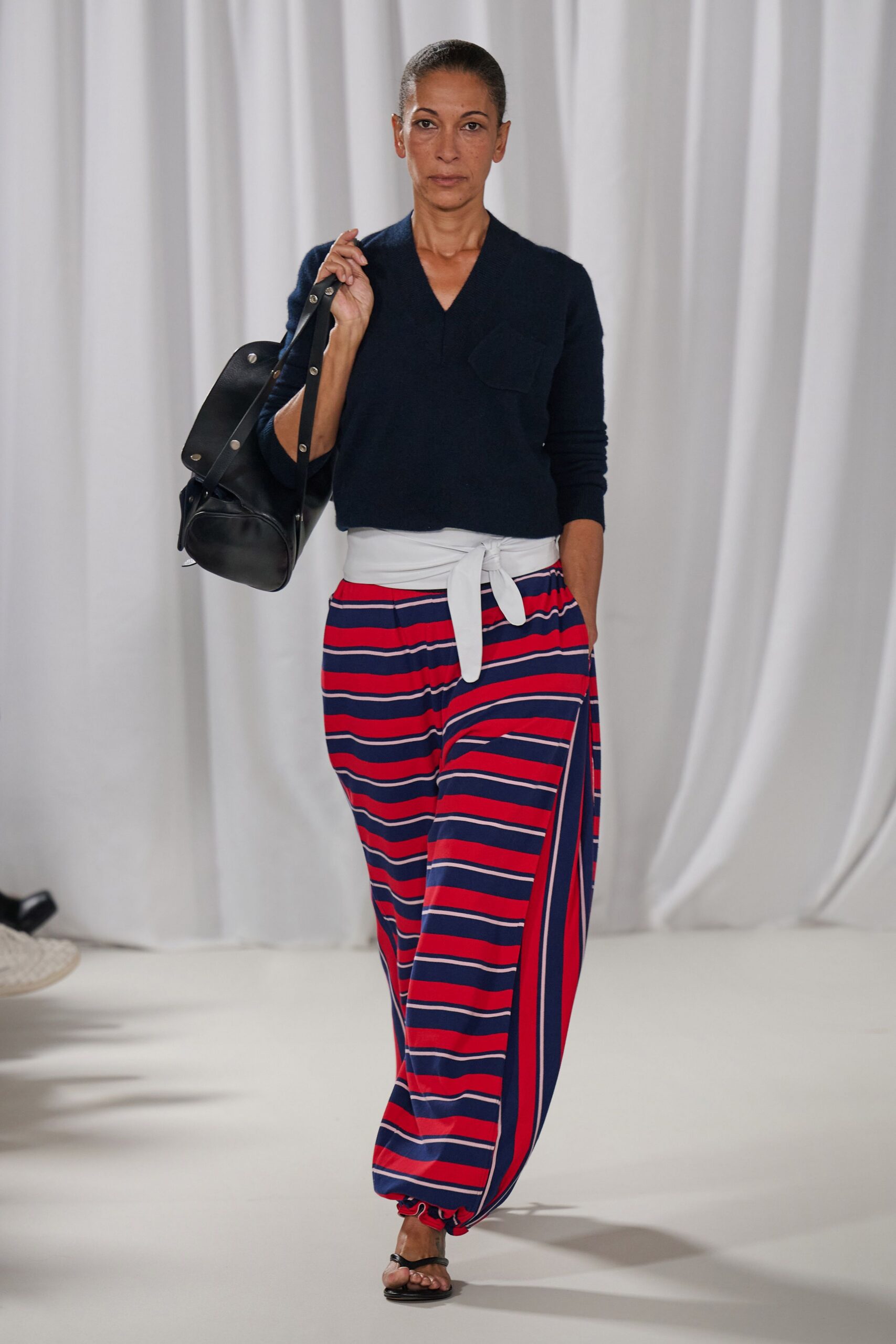 Navy fine-knit V-neck sweater with a single patch pocket. Red and navy striped wide trousers, cinched with a white self-tie wrap waistband. Black leather duffel with silver studs along the strap. Black thong sandals, flat and barely there. Walking from the ferry with a weekender and salt still drying on your skin.