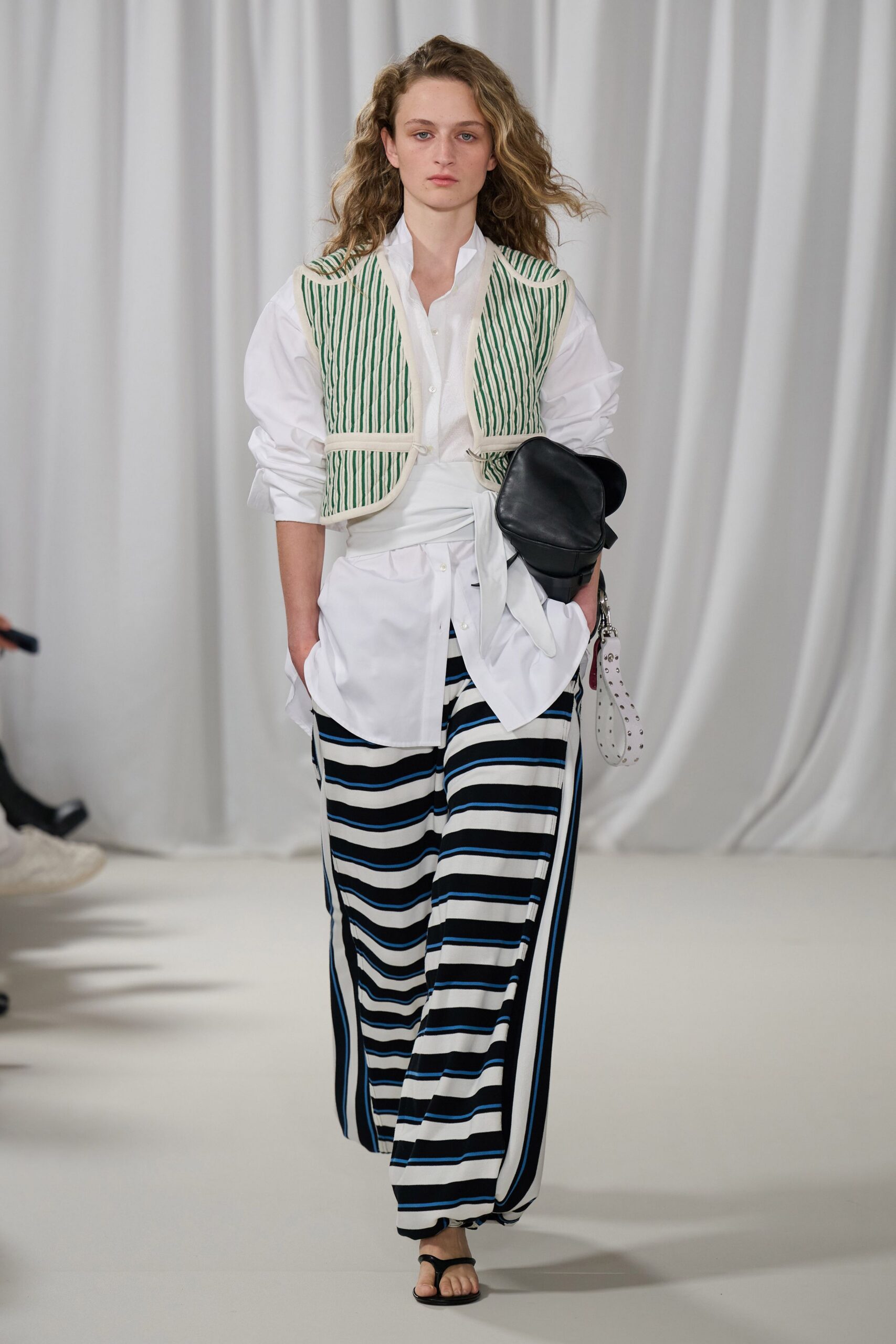 Cream cropped vest with narrow green vertical quilting and curved binding. White poplin shirt, collar popped and waist knotted askew. Wide striped trousers in black, white and cobalt jersey. Black leather pouch with a white studded wrist loop. Black thong sandals. Walking to the harbor café before the parasols are opened.