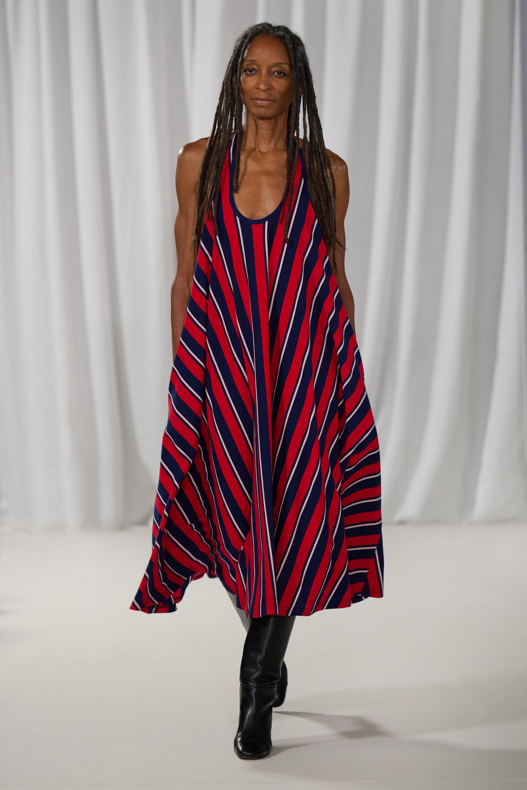 Swingy tent dress in red and navy jersey, striped on the bias. Black knee boots with softly creased shafts and squared toes. Walking across a ferry deck before the wind turns cold.