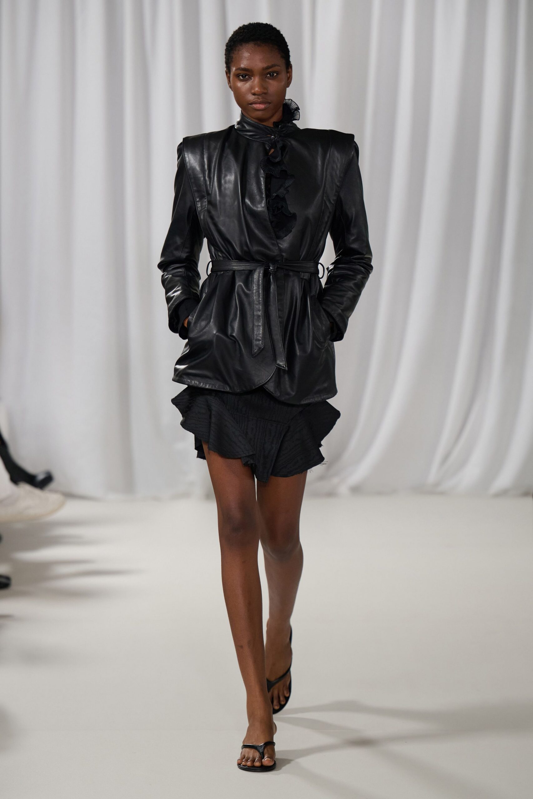 Black leather jacket with pointed shoulders and a self-tie cinched waist. Sheer black ruffle trimming the throat and peeking through the front slit. Black jacquard mini with a flounced handkerchief hem, one edge lightly frayed. Black thong-strap heels. For reading bad news from a hotel printout before breakfast service.