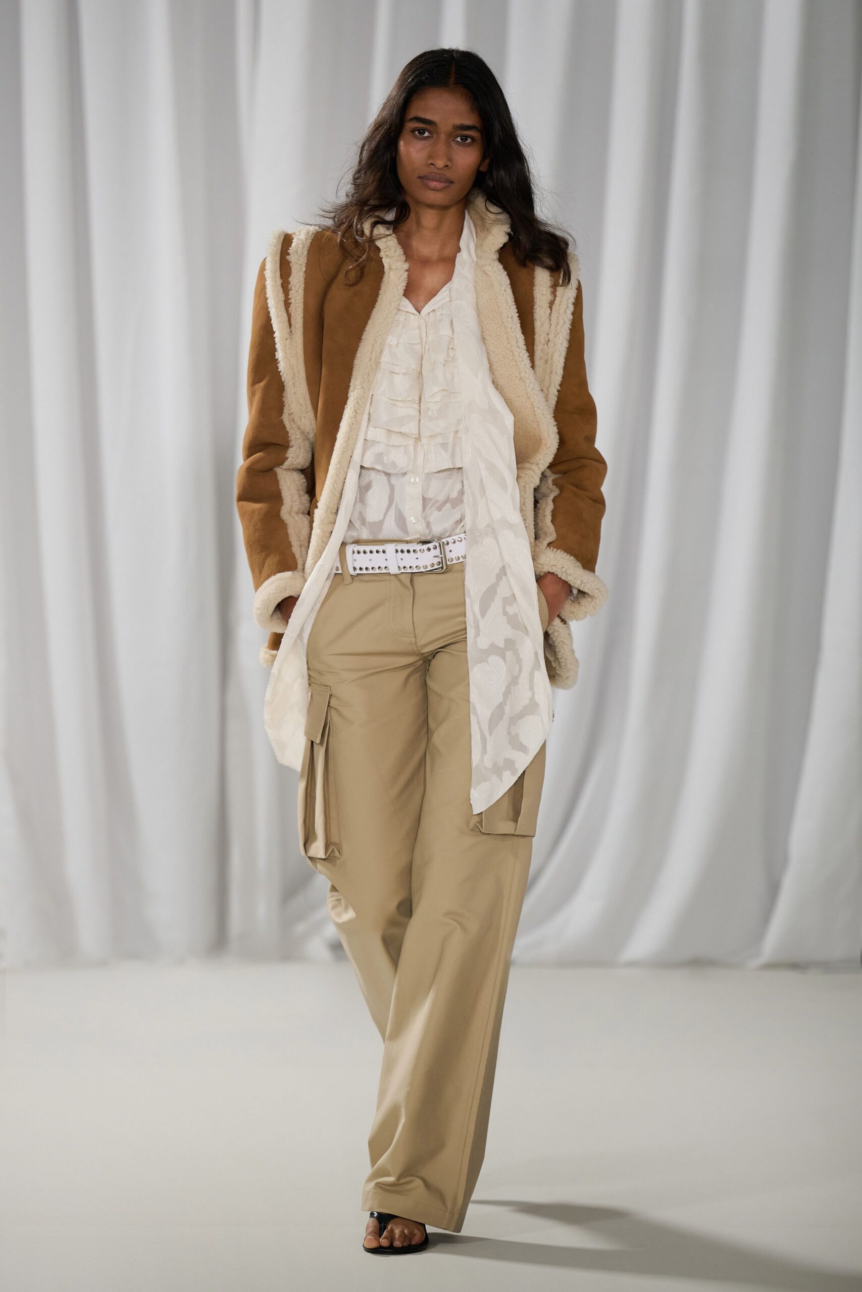 Tobacco suede jacket edged in cream shearling, seams left visibly raw. Ivory chiffon blouse with soft ruffles and a long jacquard tail. White leather belt punched all over with silver studs. Sand cargo trousers with flat side pockets at mid thigh. Black thong sandals. For the slow walk back from lunch at a desert resort.