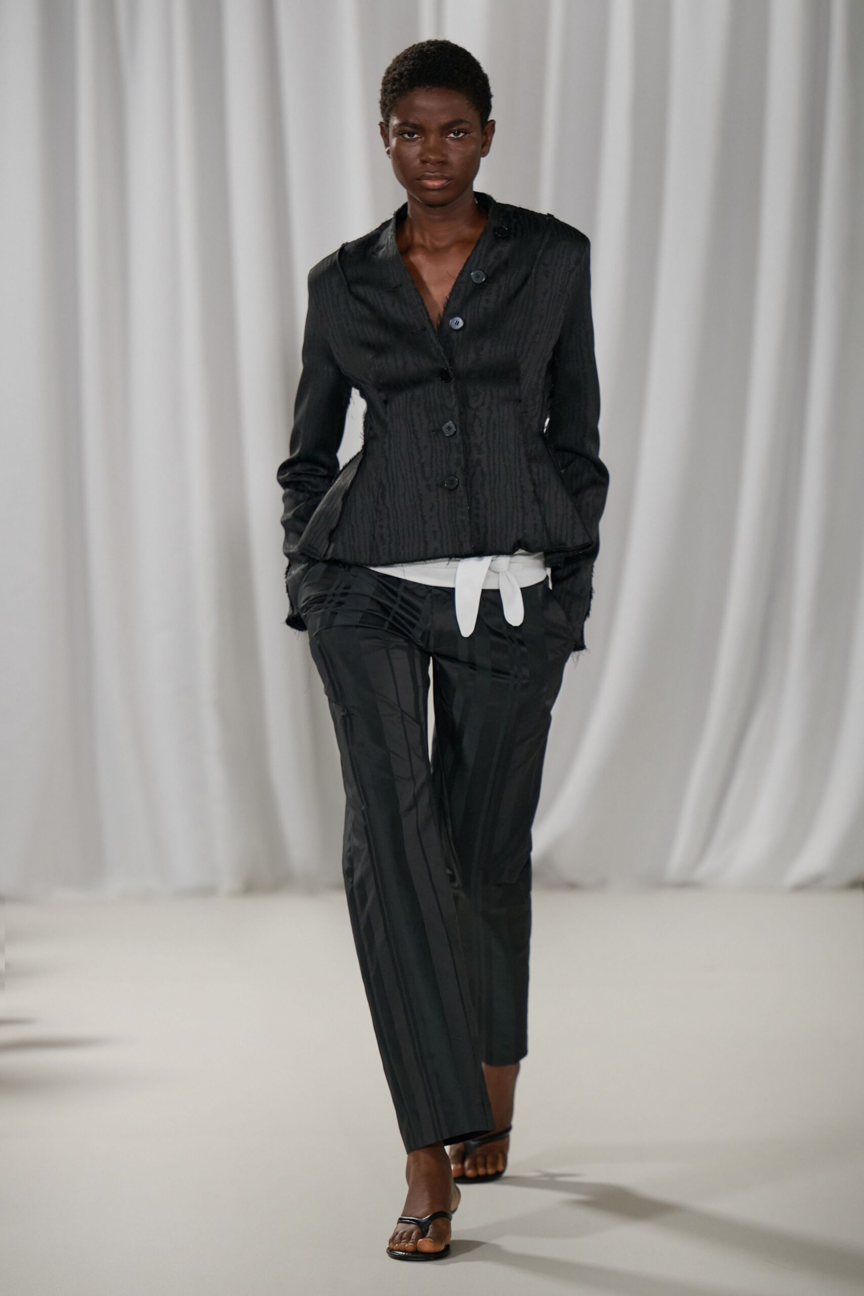 Black jacquard jacket, sharply nipped with raw edges at the cuff. White underlayer peeking out in a long tied tab. Black ankle trousers with tonal plaid sheen and a pressed crease. Barely there black thong sandals. For crossing a silent hotel lobby before breakfast service starts.