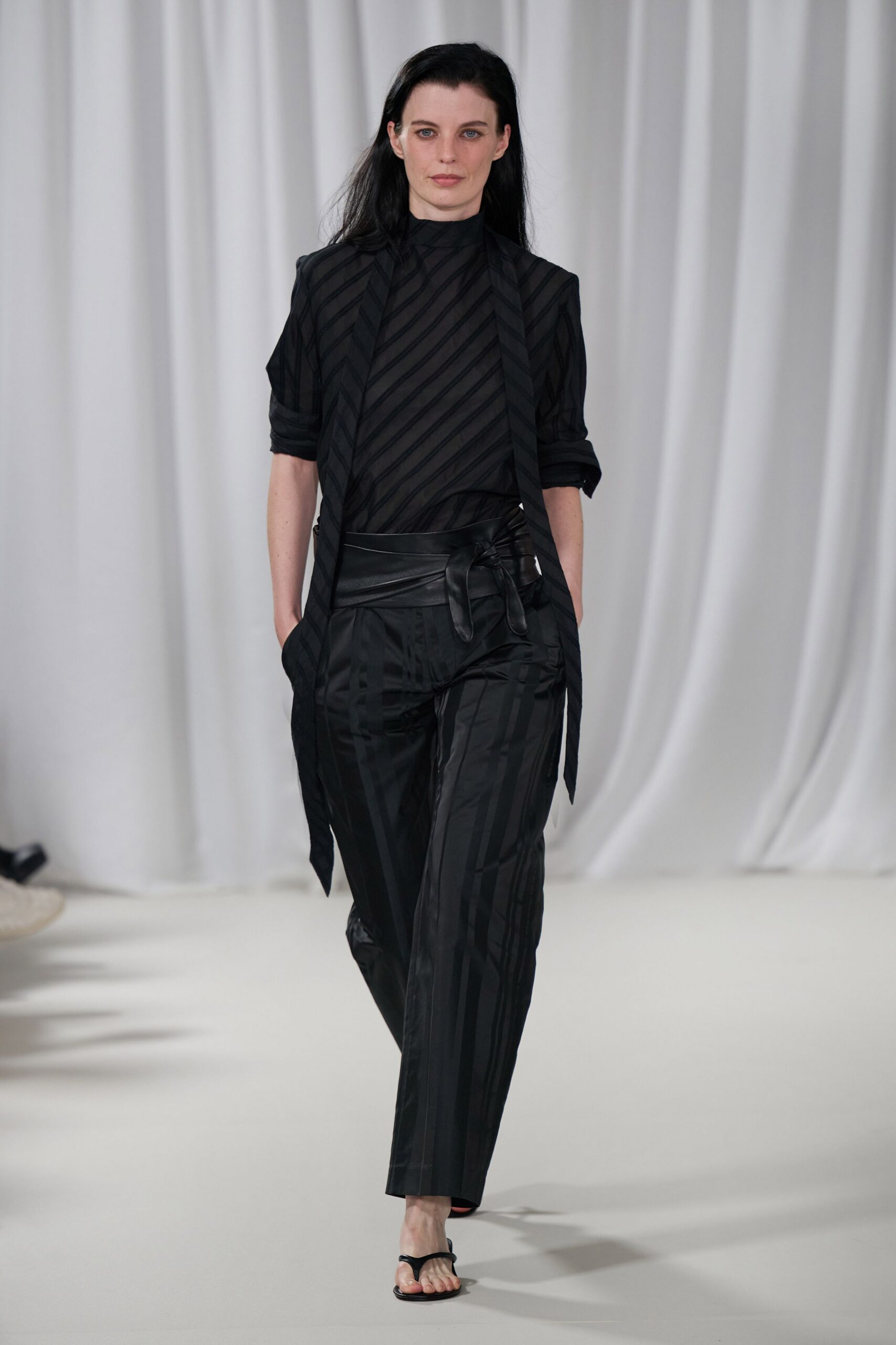 Sheer black blouse with tonal diagonal striping and a high neck. Long striped panels hanging loose past the hips. Black satin trousers with shadow stripes and a wide wrap waistband. Bare black thong sandals. For the quiet table in a hotel bar after the conference thins out.