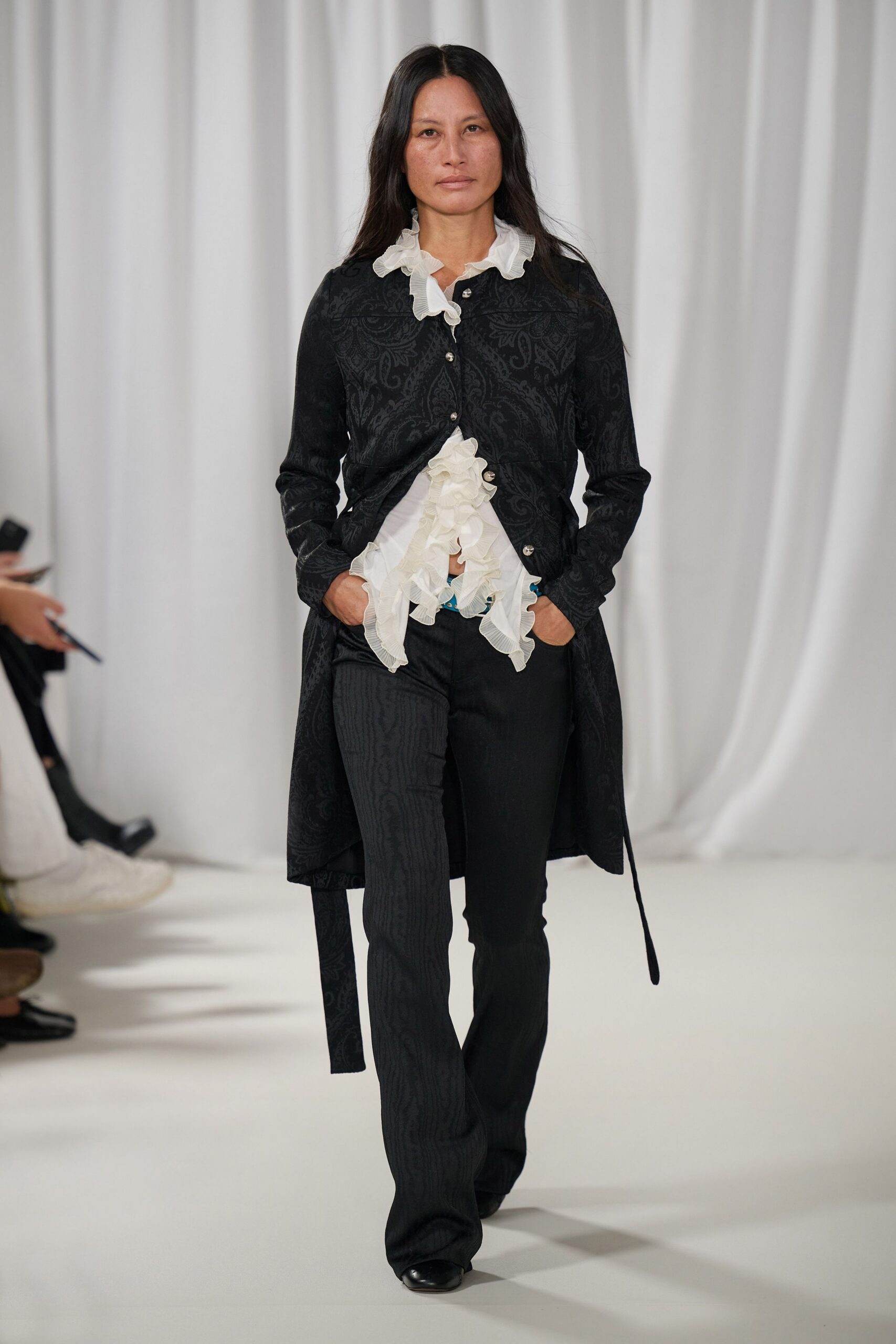White georgette blouse with dense ruffles spilling from collar to hem. Black paisley jacquard coat, pearl buttons pulling slightly at the waist. Black trousers with faint moire striping down one leg. Square-toe black leather shoes. For reading old letters in a drafty house before the radiators wake.