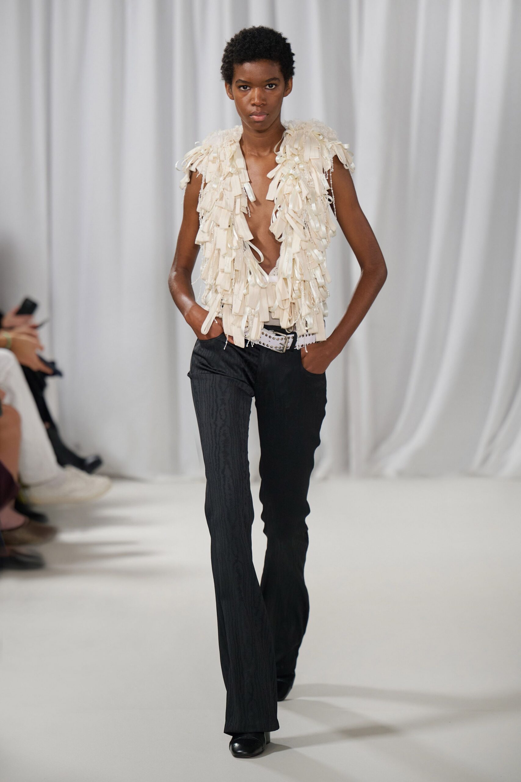 Ivory sleeveless vest covered in dangling grosgrain loops and ribbon tabs. White lace triangle bralette peeking through the plunging front. White studded belt with a square buckle, riding low on the hips. Black bootcut trousers in a faint moire stripe. For crossing a hotel lobby still wearing last night's wristband.