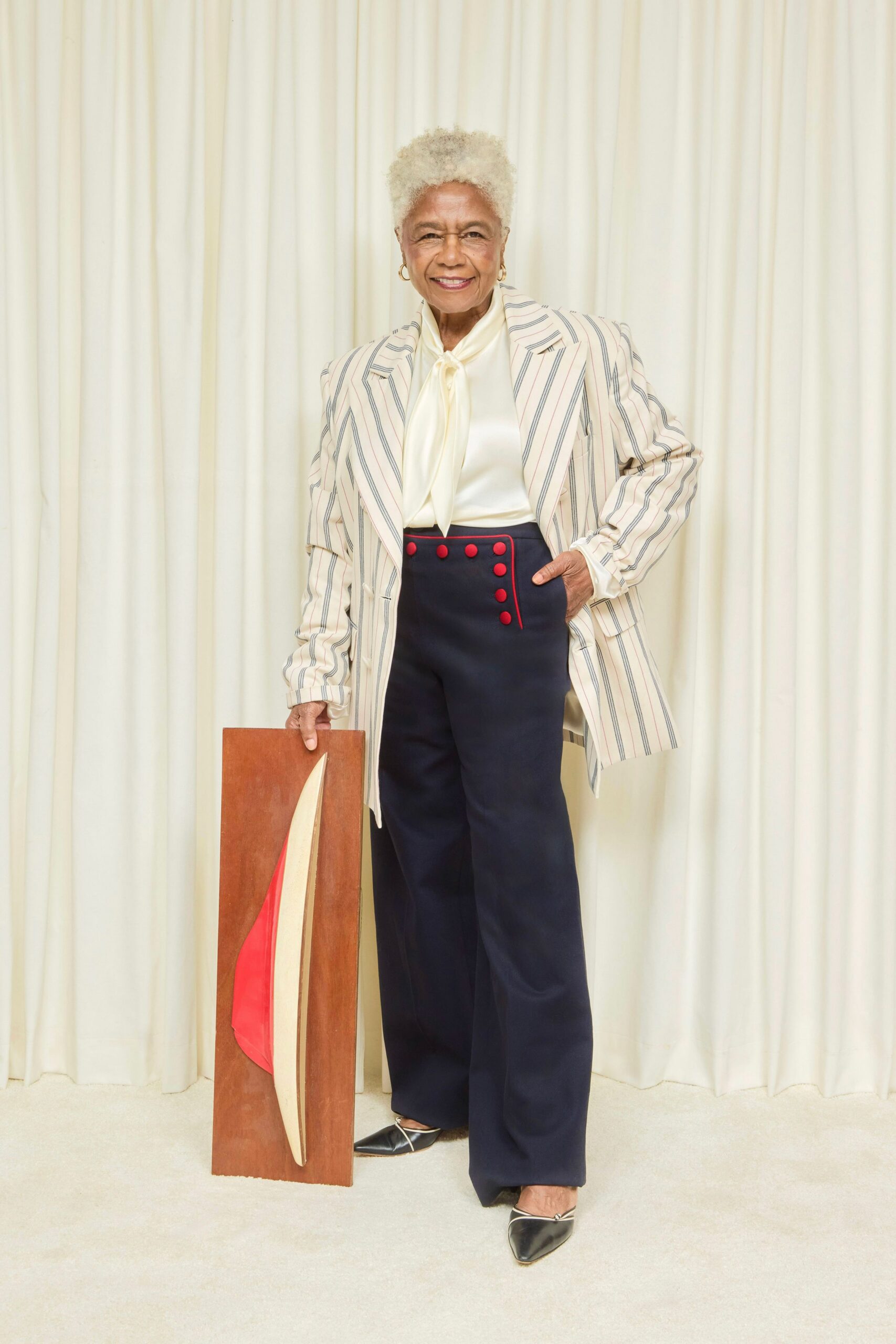 Cream blazer with spaced navy and blush pinstripes, cut long through the hip. Ivory satin tie-neck blouse, the bow falling in a loose narrow drape. Navy sailor trousers with red piping and two neat rows of covered buttons. Black pointed flats edged in white. At a small museum fundraiser, standing beside the auction sculpture.