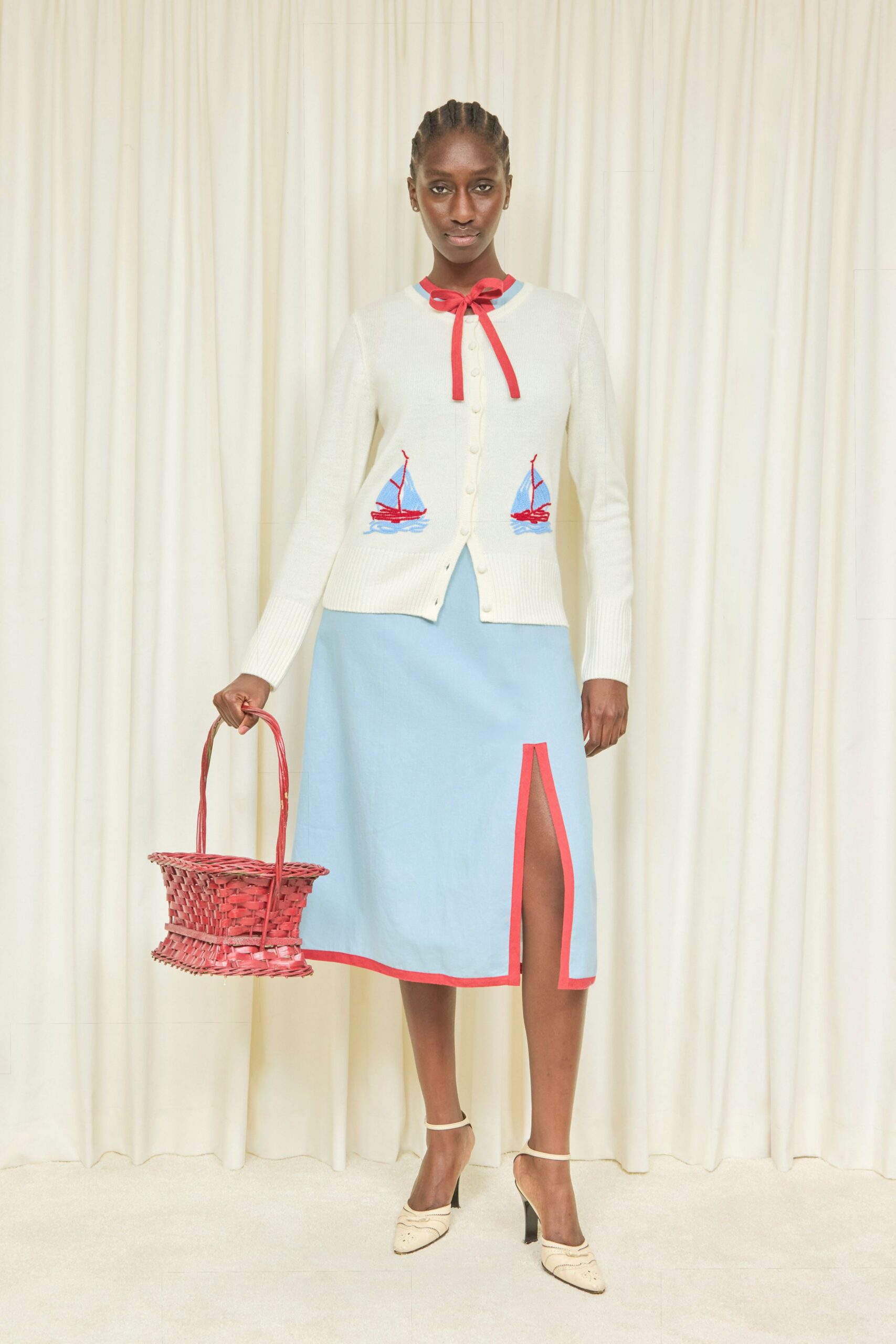 Cream fine-knit cardigan with sailboat intarsia and a bright red bow. Powder-blue midi dress with coral binding tracing the high front slit. Coral wicker basket bag, slightly pinched at the middle. Cream slingback mules with black heels and punched wavy seams. Walking back from the marina with melted sorbet on your wrist.