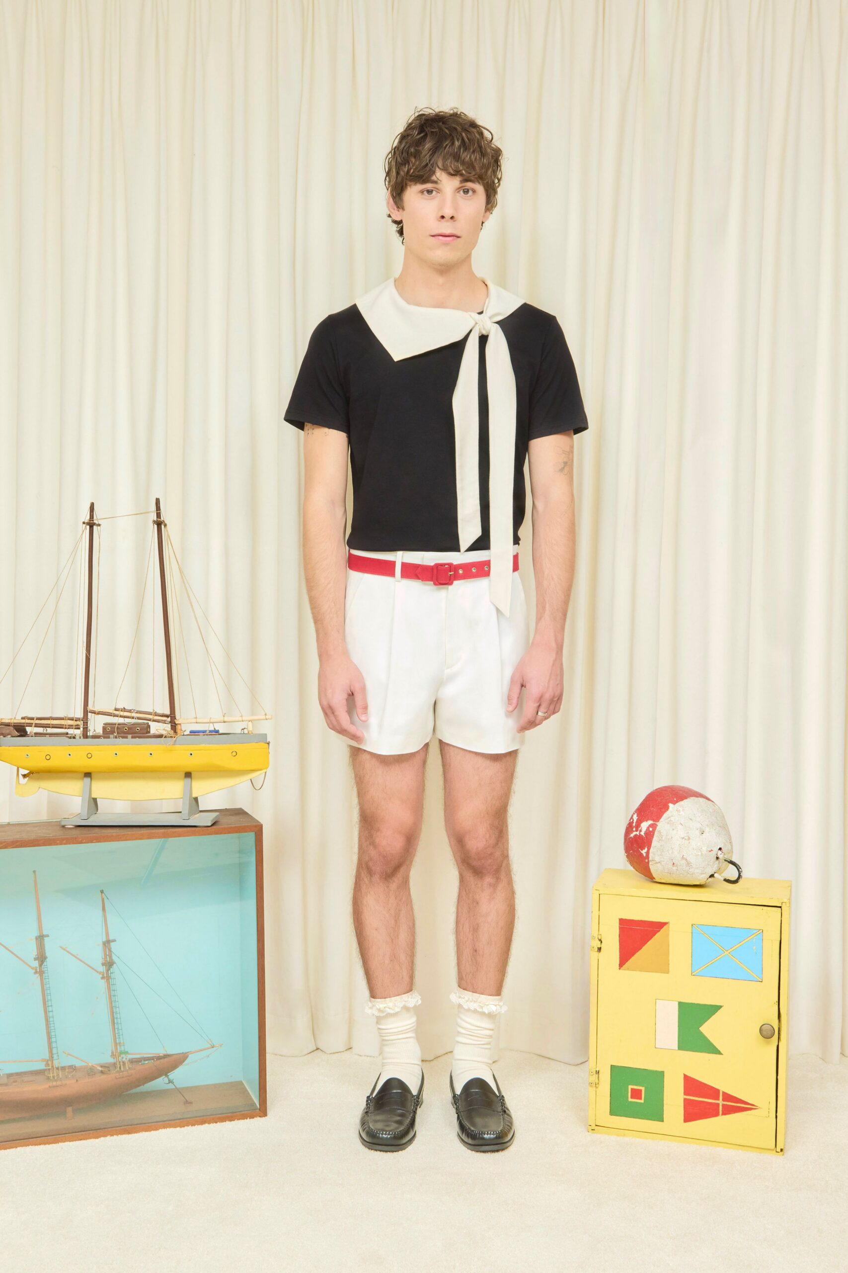 Black knit tee with a cream sailor scarf tied off-center. White tailored shorts with a clean pressed front. Coral belt with a small covered buckle. White socks, ruffled elastic peeking above the ankle. Black leather penny loafers with raised apron stitching. For a harbor lunch after buying postcards at the marina shop.