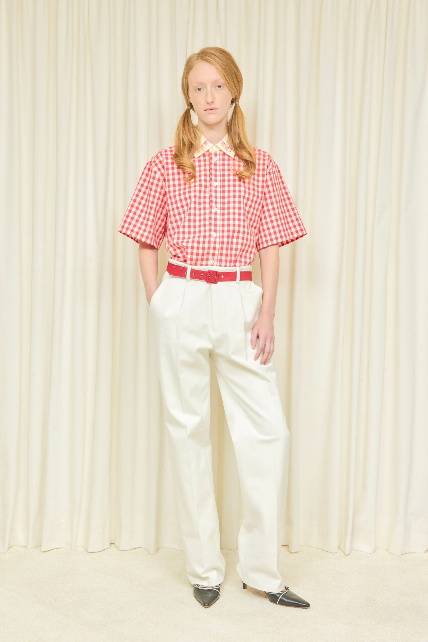 Red-and-white gingham shirt with wide elbow sleeves and a cream collar tip. Cherry red belt with small brass grommets and a covered buckle. High-waisted cream trousers, sharply pressed and pooling lightly at the shoe. Black pointed mules traced with a thin white piping. Sunday open house, someone cut peonies for the hall table.
