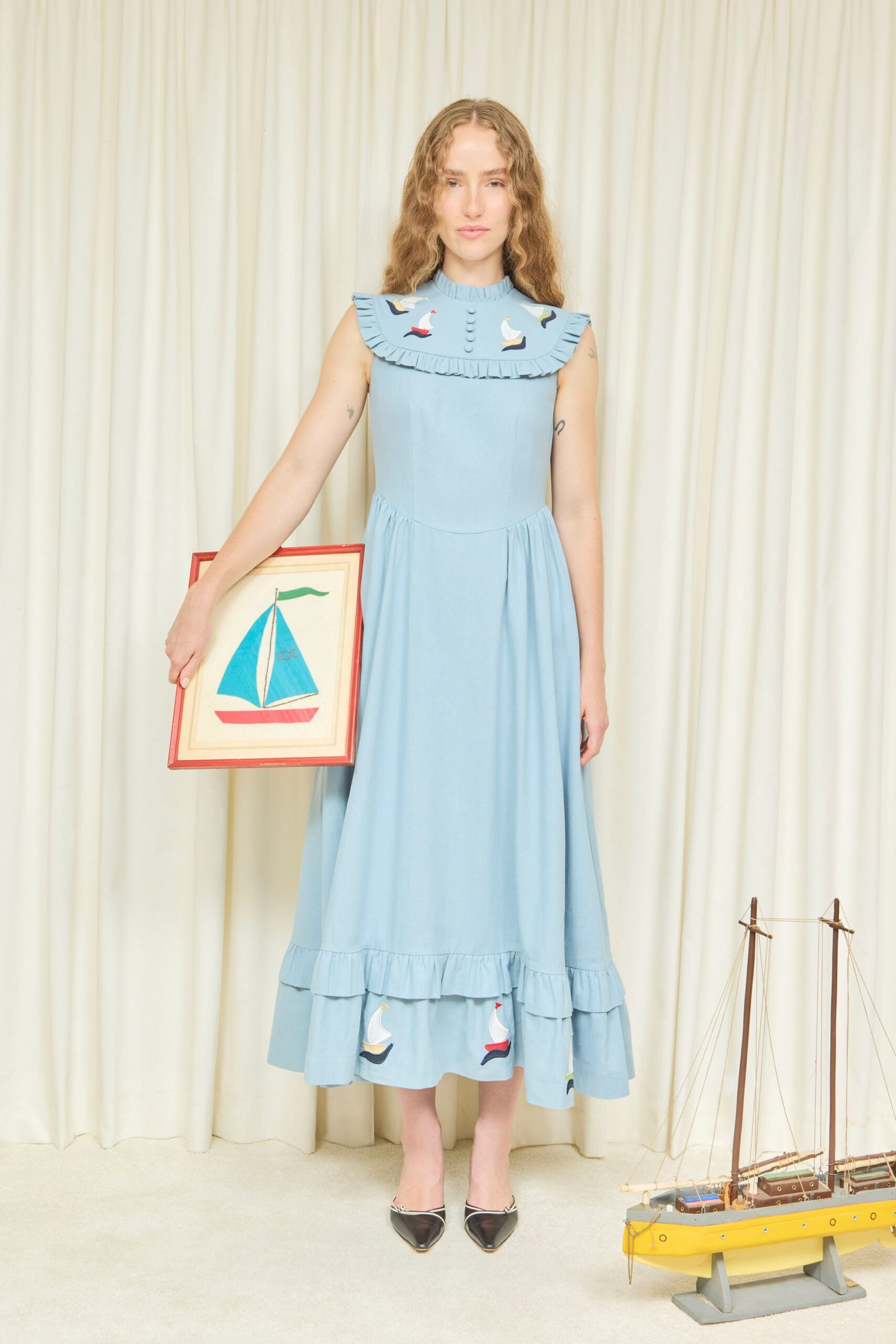 Powder-blue sleeveless dress with a broad ruffled bib, pintucked buttons and appliqued sailboats. Black pointed mules edged in silver at the topline. For hanging a child's boat painting in the summer house hallway.