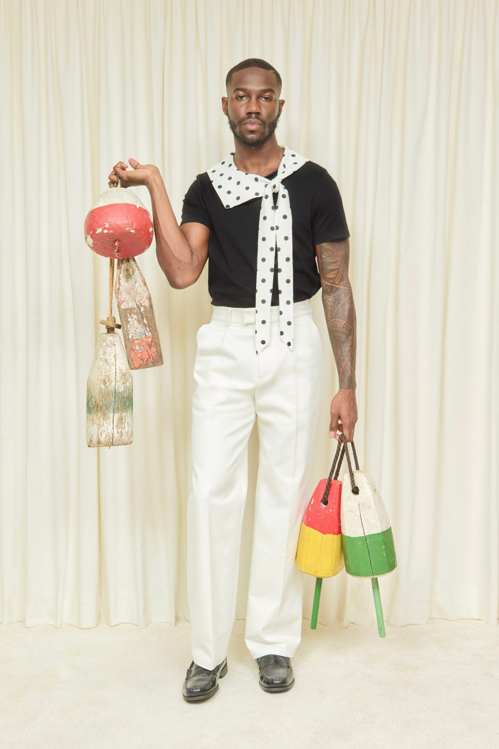 Black jersey tee with a plain close crewneck. White polka-dot scarf collar tied long down the chest. High-waisted white trousers with a hard front crease. Black leather loafers with apron stitching at the toe. Weathered painted fishing buoys carried by rope loops. Walking back from the dock in borrowed dinner clothes.