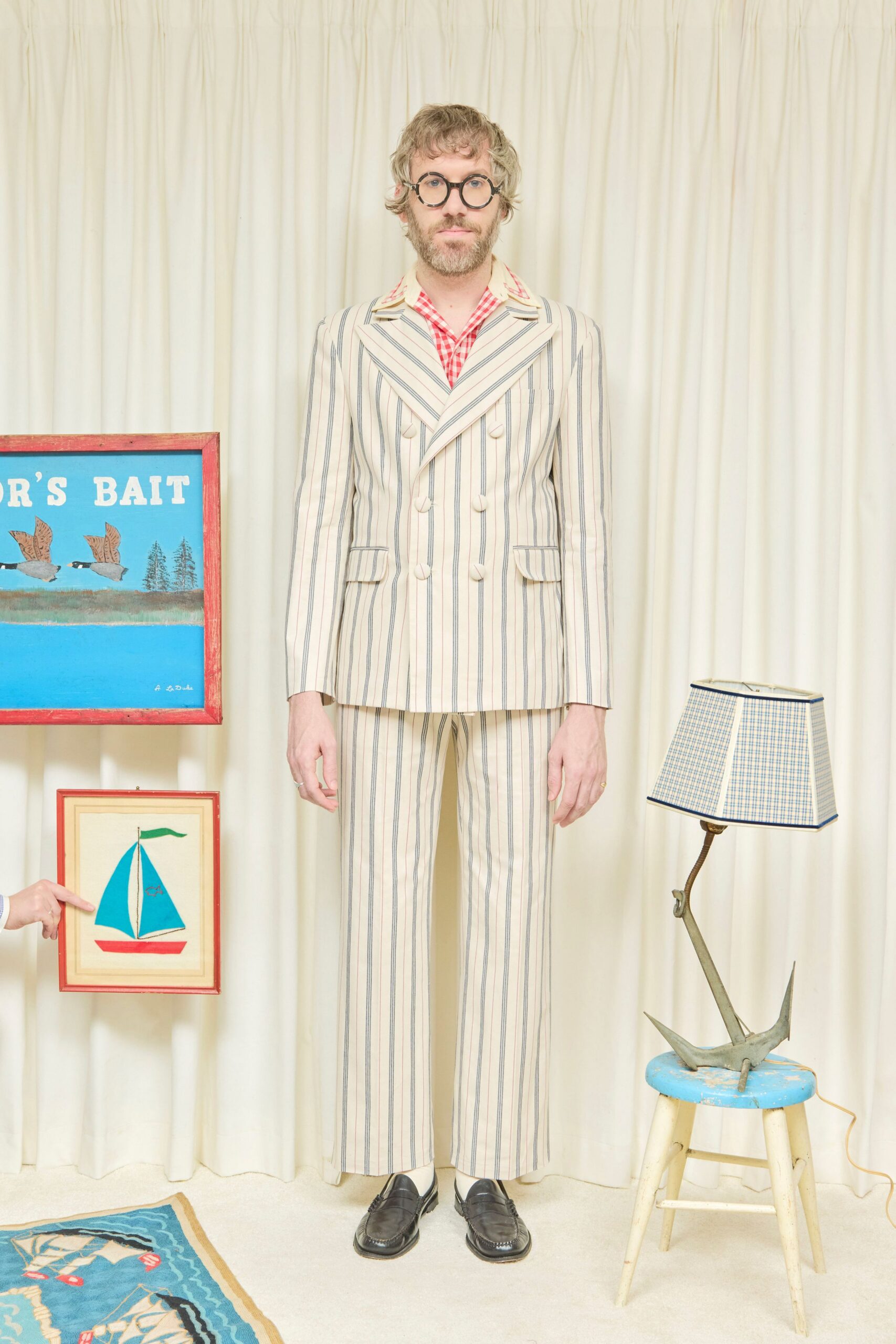 Round black frames with narrow silver hinges. Cream double-breasted blazer in blue pencil stripes, peaked lapels cut wide. Red gingham shirt open at the throat. Matching striped trousers with a long easy break. Black leather penny loafers with white socks showing. For hanging a crooked sailboat print in a rented beach cottage.