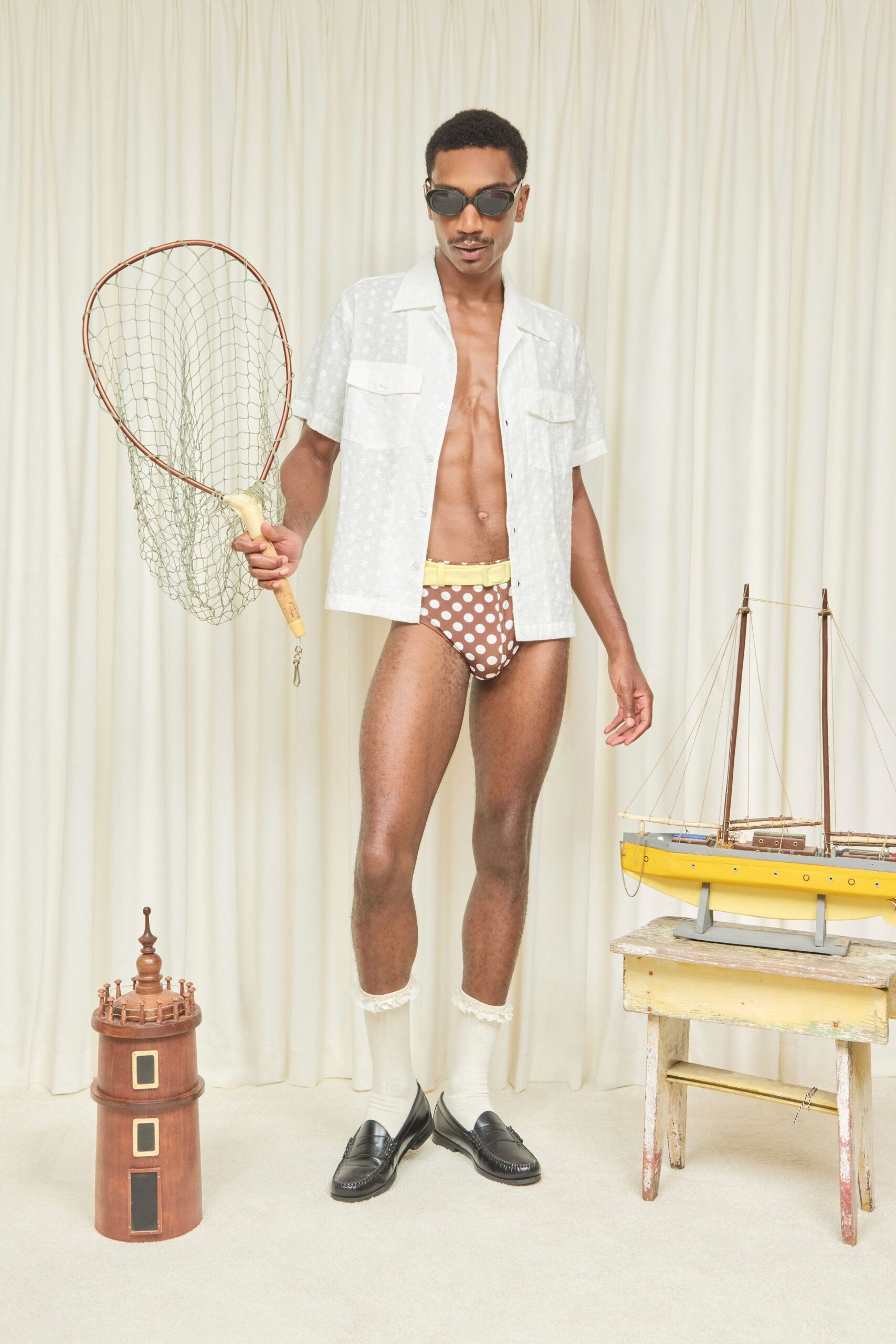 Small black oval frames. Sheer white camp-collar shirt with faint woven flowers and twin flap pockets. Brown briefs dotted in white, topped with a pale yellow belt. White knee socks trimmed with ruffled lace. Black leather penny loafers with raised apron stitching. For checking crab traps after lunch at a rented beach house.