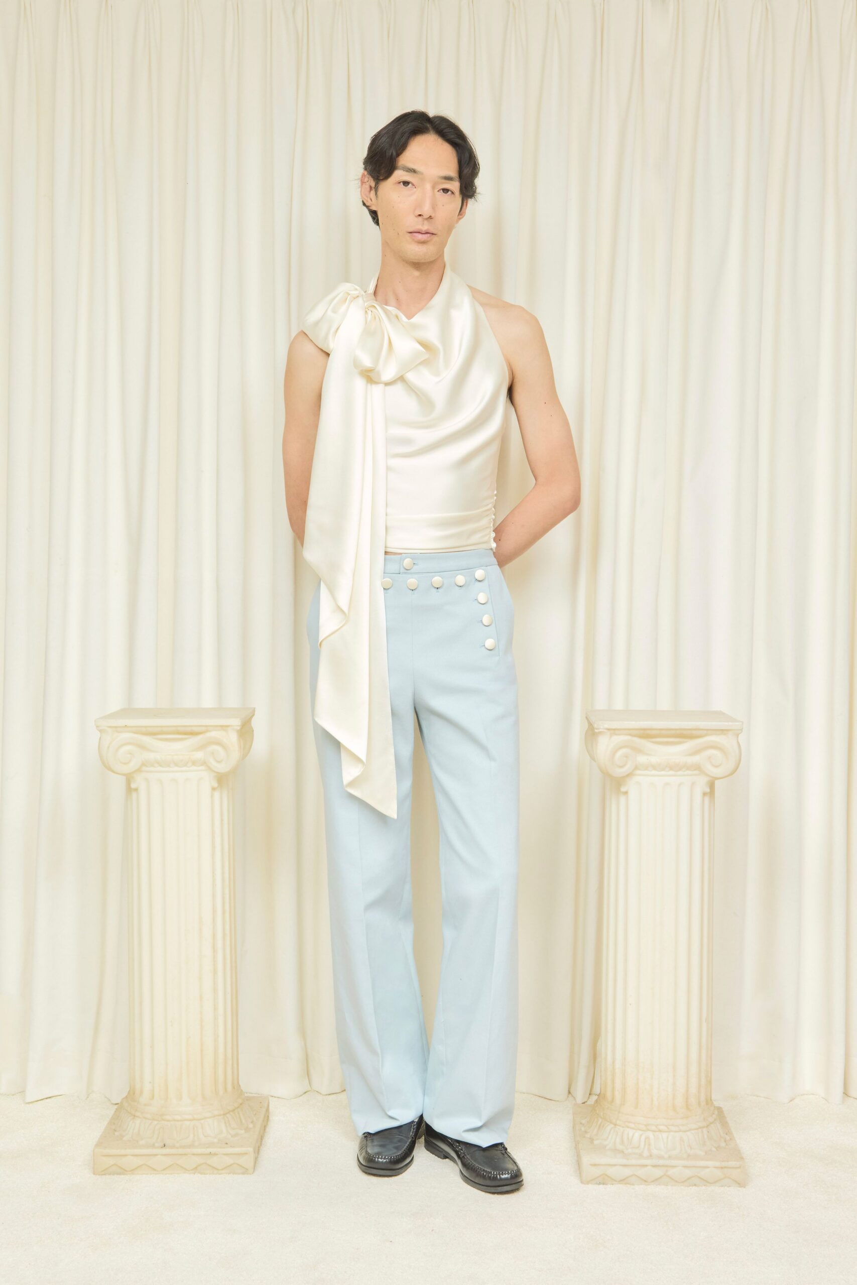 Ivory satin one-shoulder top, knotted high with a long scarf drape. Powder-blue flared trousers, waistband tipped with a row of white dome buttons. Black leather loafers with raised apron stitching. Under painted columns after the wedding, before anyone finds the cake knife.