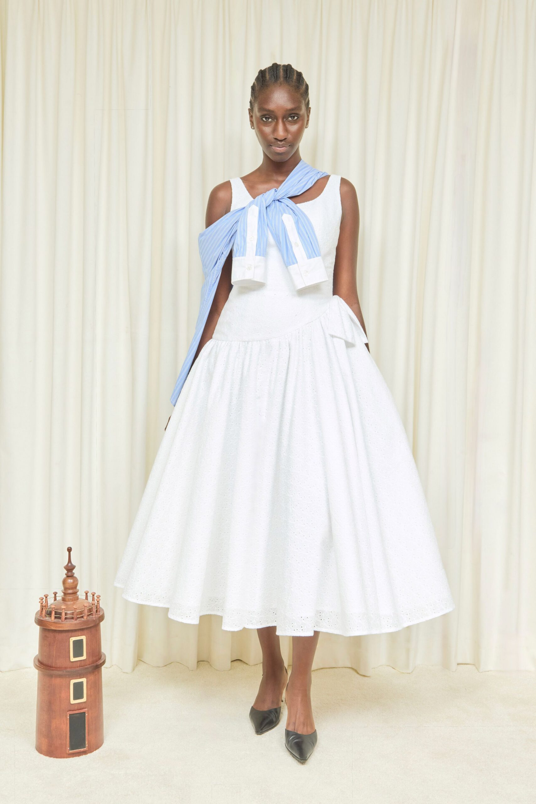 White broderie anglaise dress with a dropped waist and wide gathered skirt. Blue banker-stripe shirt knotted through the neckline, cuffs hanging unevenly. Black leather pumps with narrow pointed toes. For a garden recital where someone dragged dining-room clothes onto the lawn.