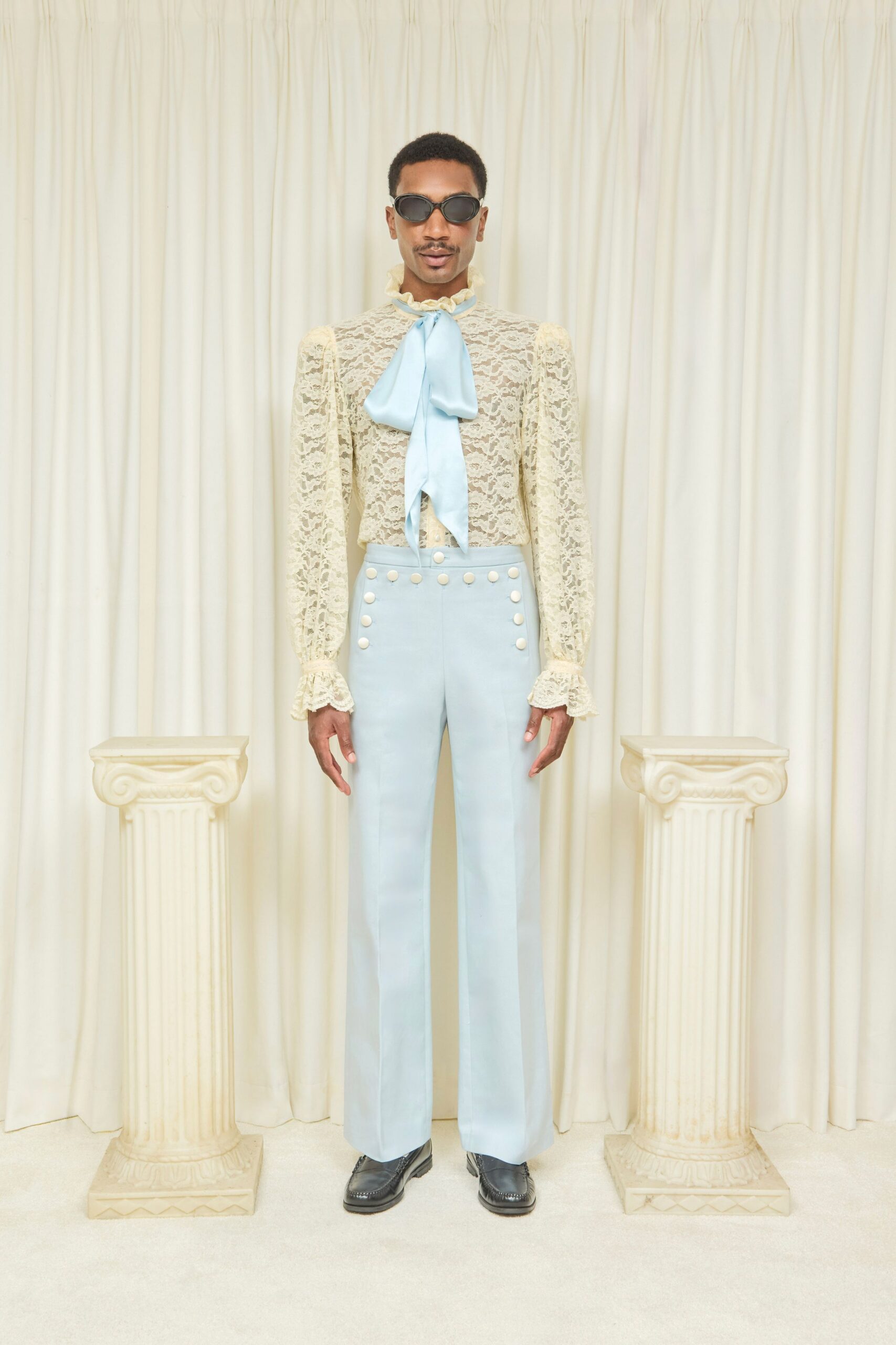Black oval sunglasses with thick glossy rims. Cream floral lace blouse, ruffled collar and cuffs, powder-blue pussy bow. Powder-blue high-waisted flares with two rows of pearl-like dome buttons. Black leather loafers with square toes and apron stitching. Sunday house tour, someone opens the curtains on a faded ballroom.