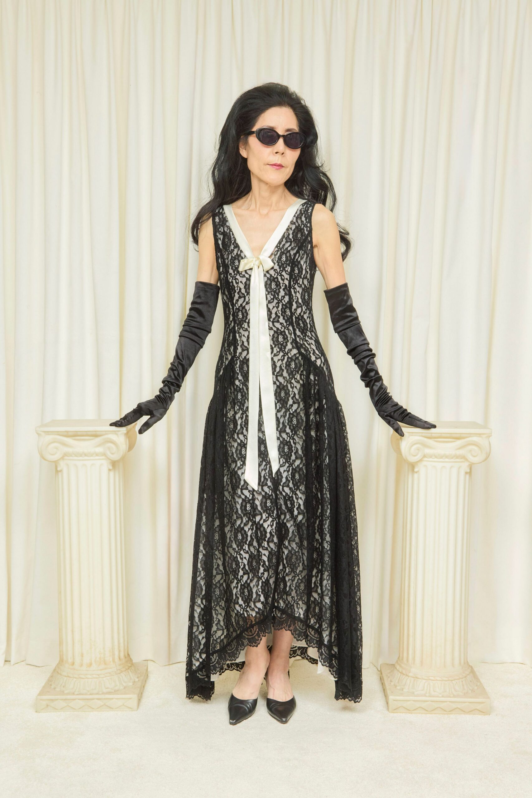 Narrow black oval frames with smoke-dark lenses. Black floral lace gown over a pale slip, finished with a long ivory bow. Opera-length black satin gloves, softly ruched at the forearm. Black leather mules with a sharp pointed toe. Hotel wedding at noon, still wearing last night's eyeliner.