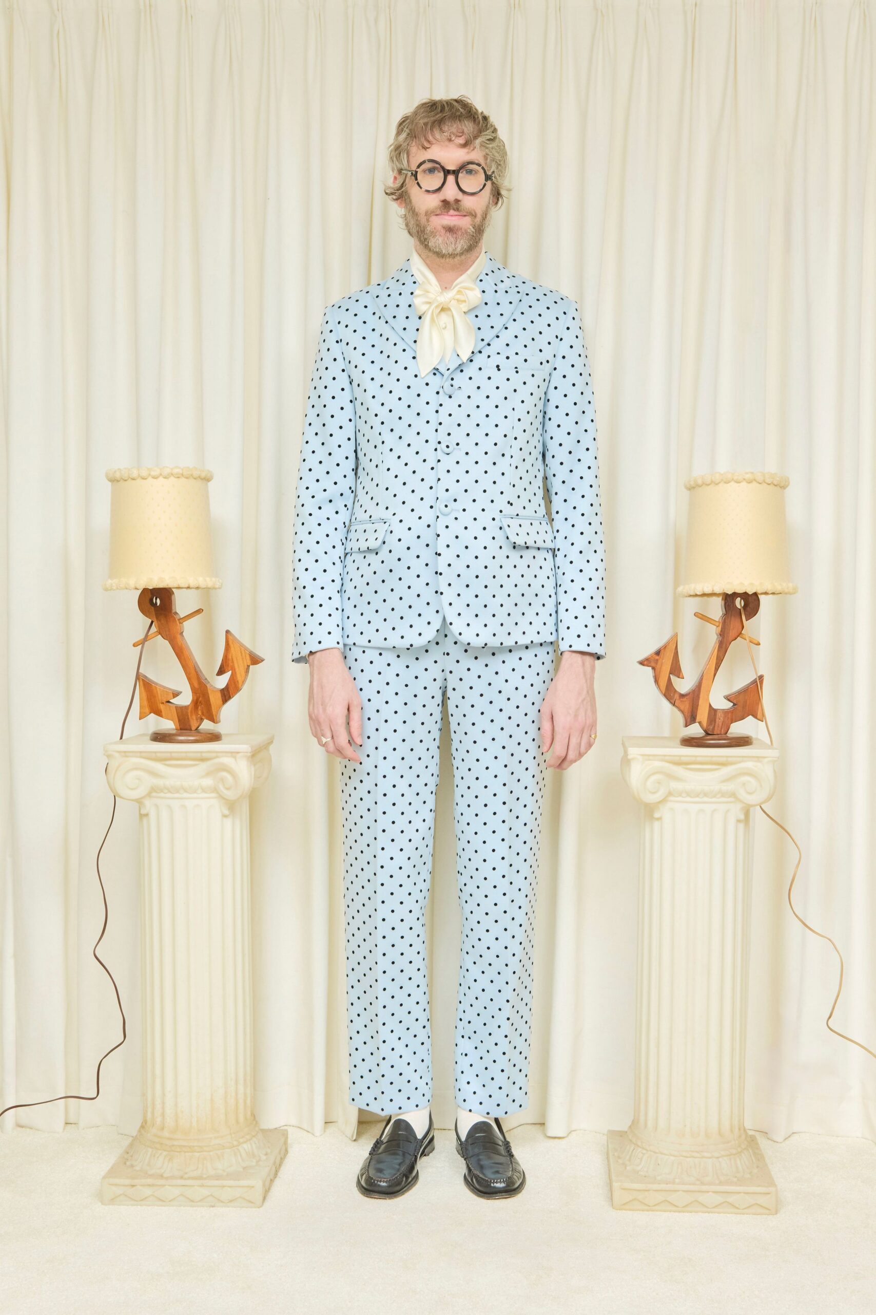 Round black frames, thick as toy spectacles. Cream satin scarf tied in a soft oversized bow. Powder-blue suit jacket dotted in black, with flap pockets. Matching slim trousers cropped just above the ankle. Black leather penny loafers with creased vamps. At a seaside wedding where the rental villa still smells faintly of bleach.