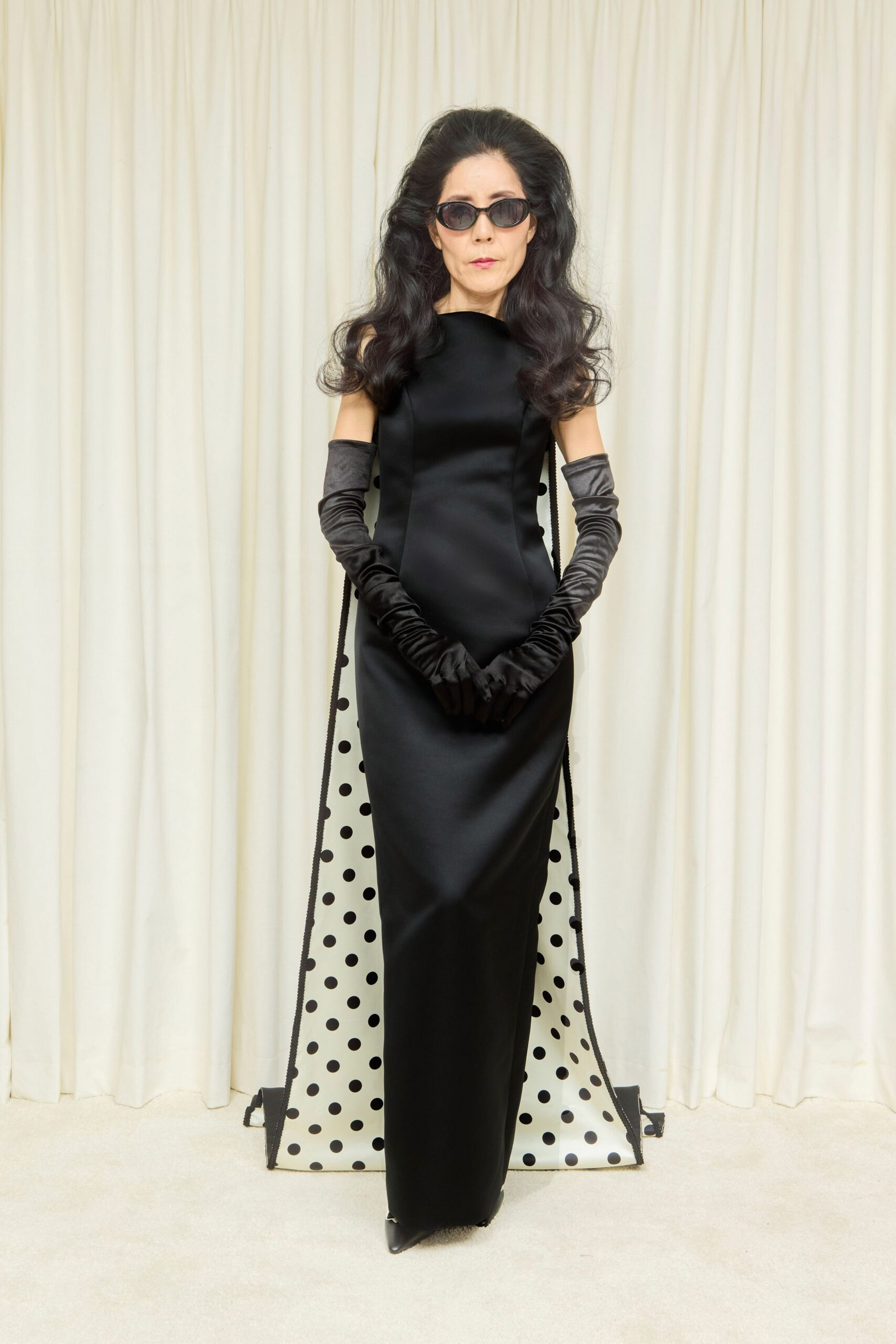 Narrow smoked oval frames. Black satin column gown with a bateau neck and side cutouts. White train scattered with black polka dots, corded in black. Gunmetal satin opera gloves, softly ruched at the wrists. Black pointed toes peeking under the hem. Hotel ballroom at noon, after the string quartet has packed up.