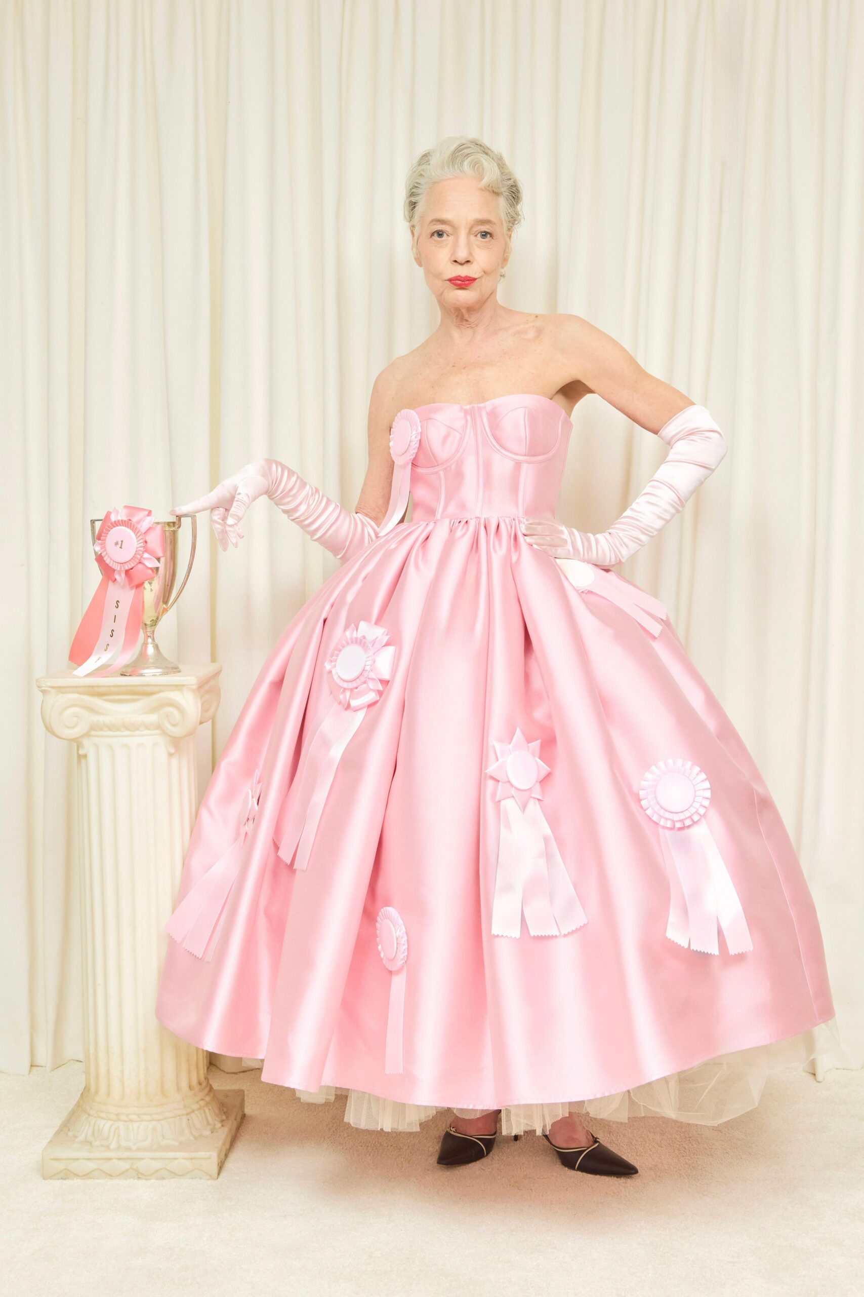 Strapless pink satin ballgown with corseted cups and prize rosettes scattered wide. Opera gloves in pale satin, ruched along the forearms. White tulle peeking below the hem at the front. Black pointed pumps with a fine pale binding at the edge. County-show prizegiving, posed beside the silver cup after everyone has gone.